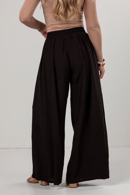 A woman shown from the back wearing black wide-leg pants with an elastic waistband, paired with a beige long-sleeve top, against a plain light gray studio background.