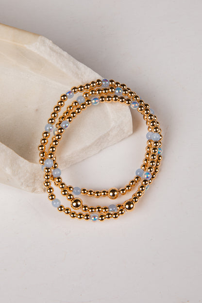 A stack of three beaded stretch bracelets with metallic gold beads and light blue faceted beads, displayed on a neutral cream-colored stone prop against a light background.