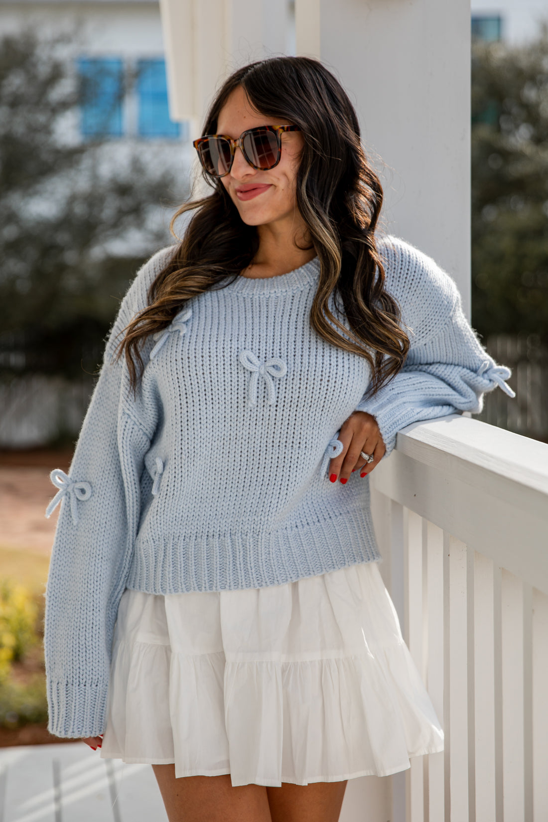 Woman wearing a light blue sweater with bow details and a white skirt on a porch.