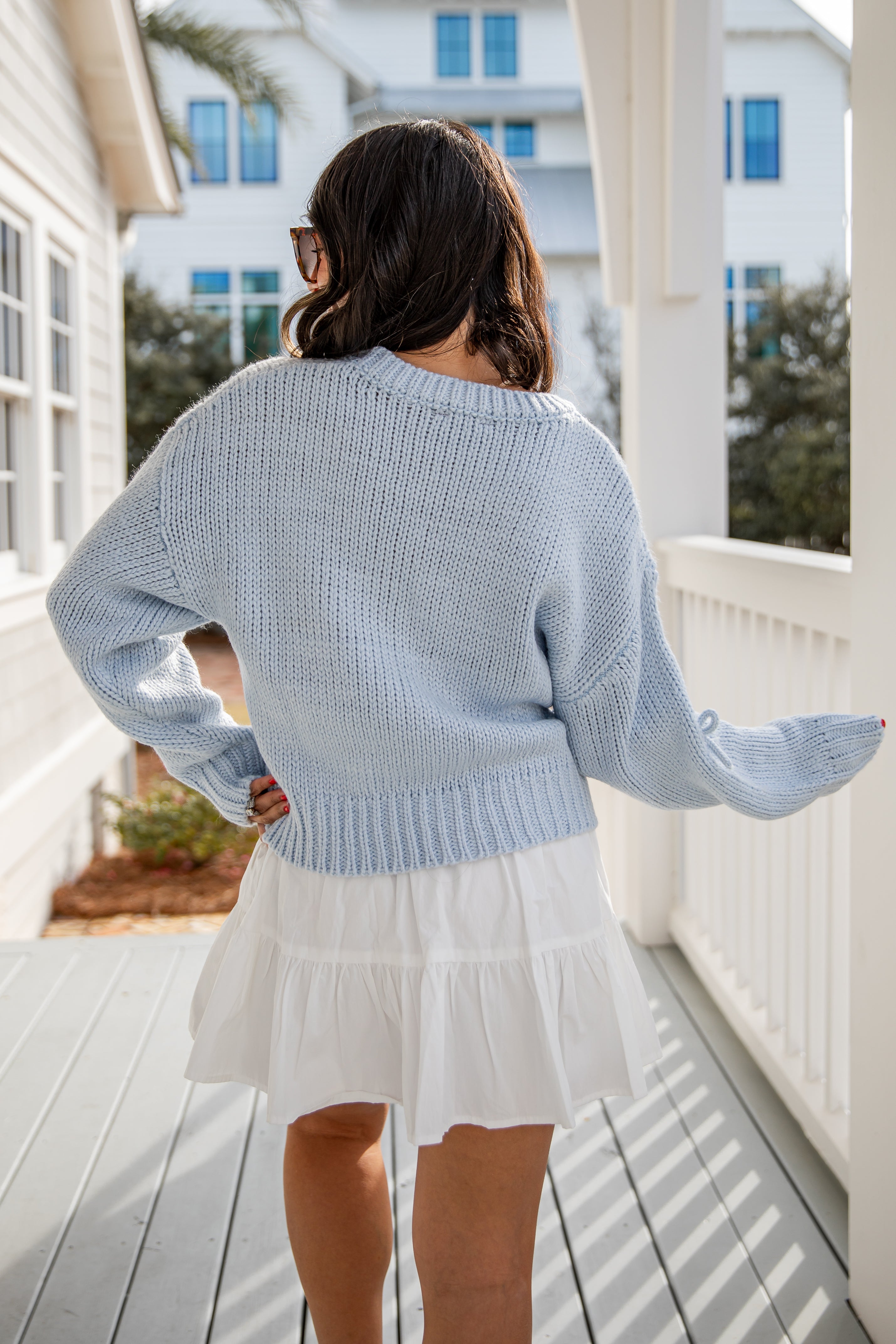 Woman wearing a light blue sweater and white skirt on a porch.