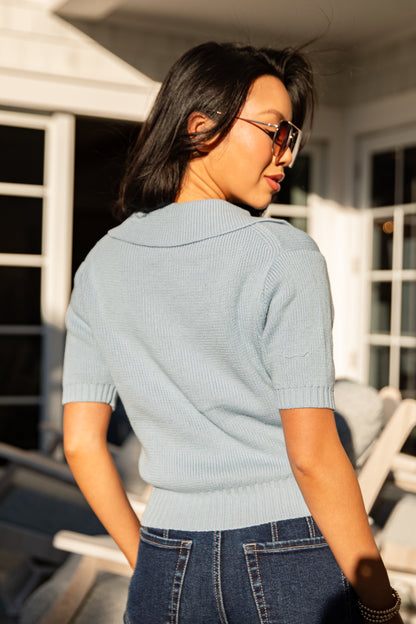 Woman wearing a light blue sweater and sunglasses, standing outdoors.