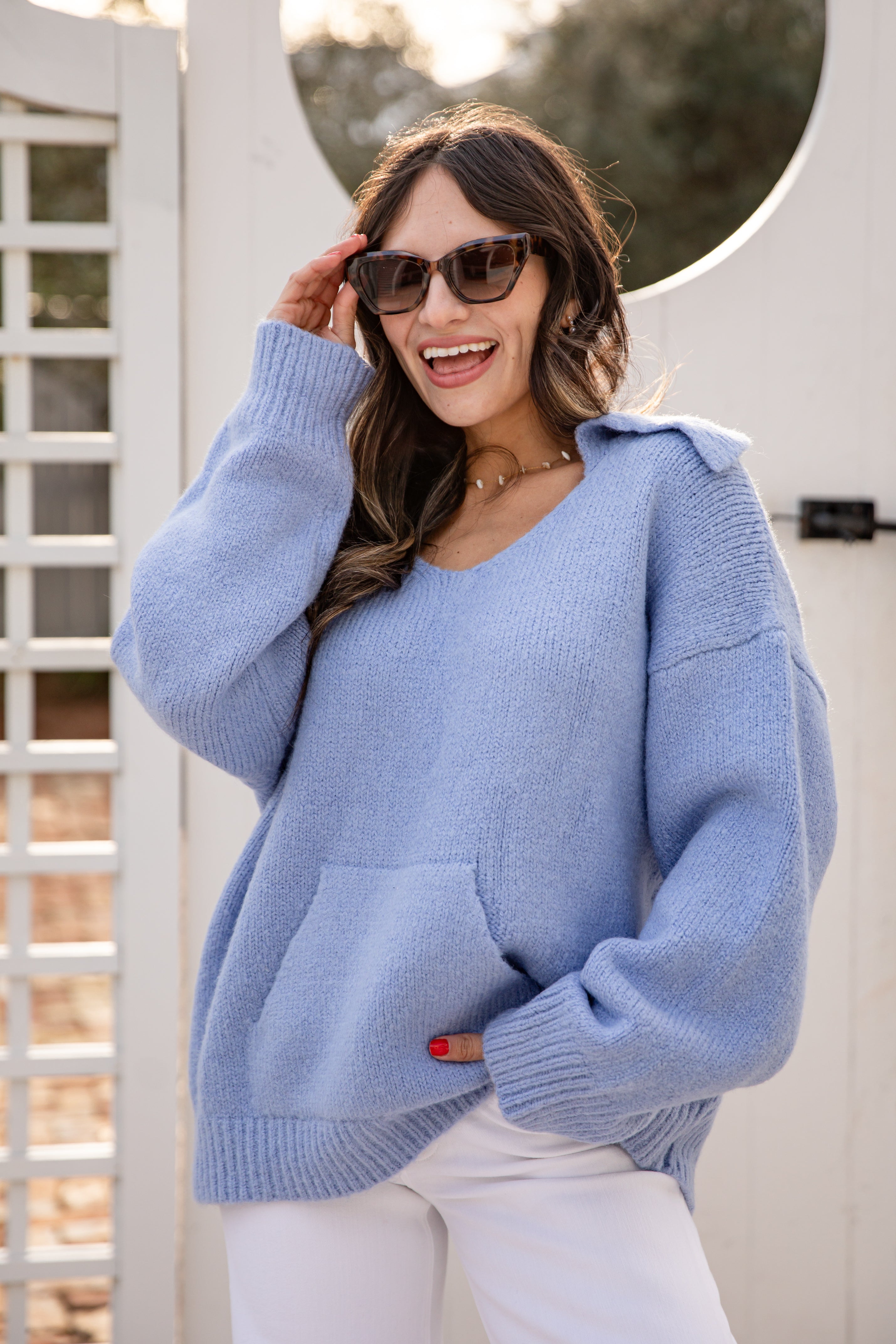 Woman wearing a light blue sweater and sunglasses, standing in front of a white door.