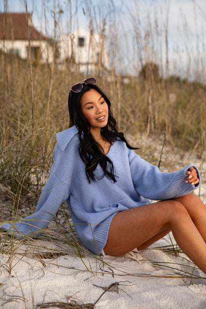 Woman in a light blue sweater sitting on sand with grass and a house in the background