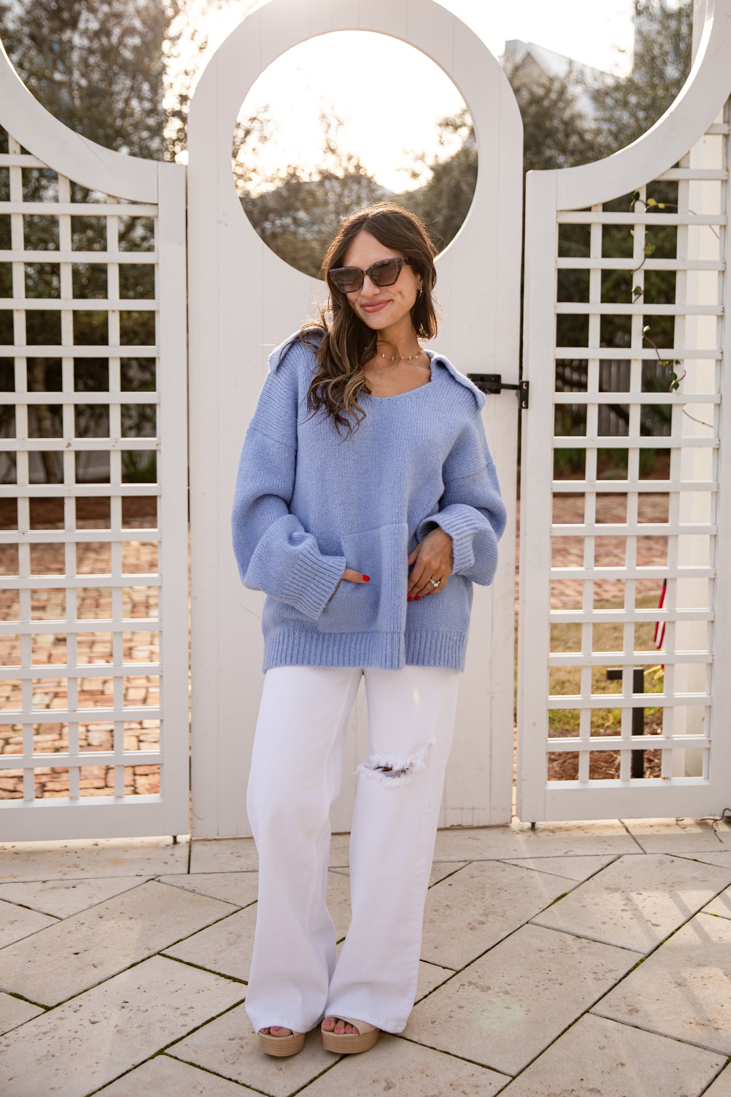 Woman wearing a blue sweater and white pants standing in front of a white lattice gate.
