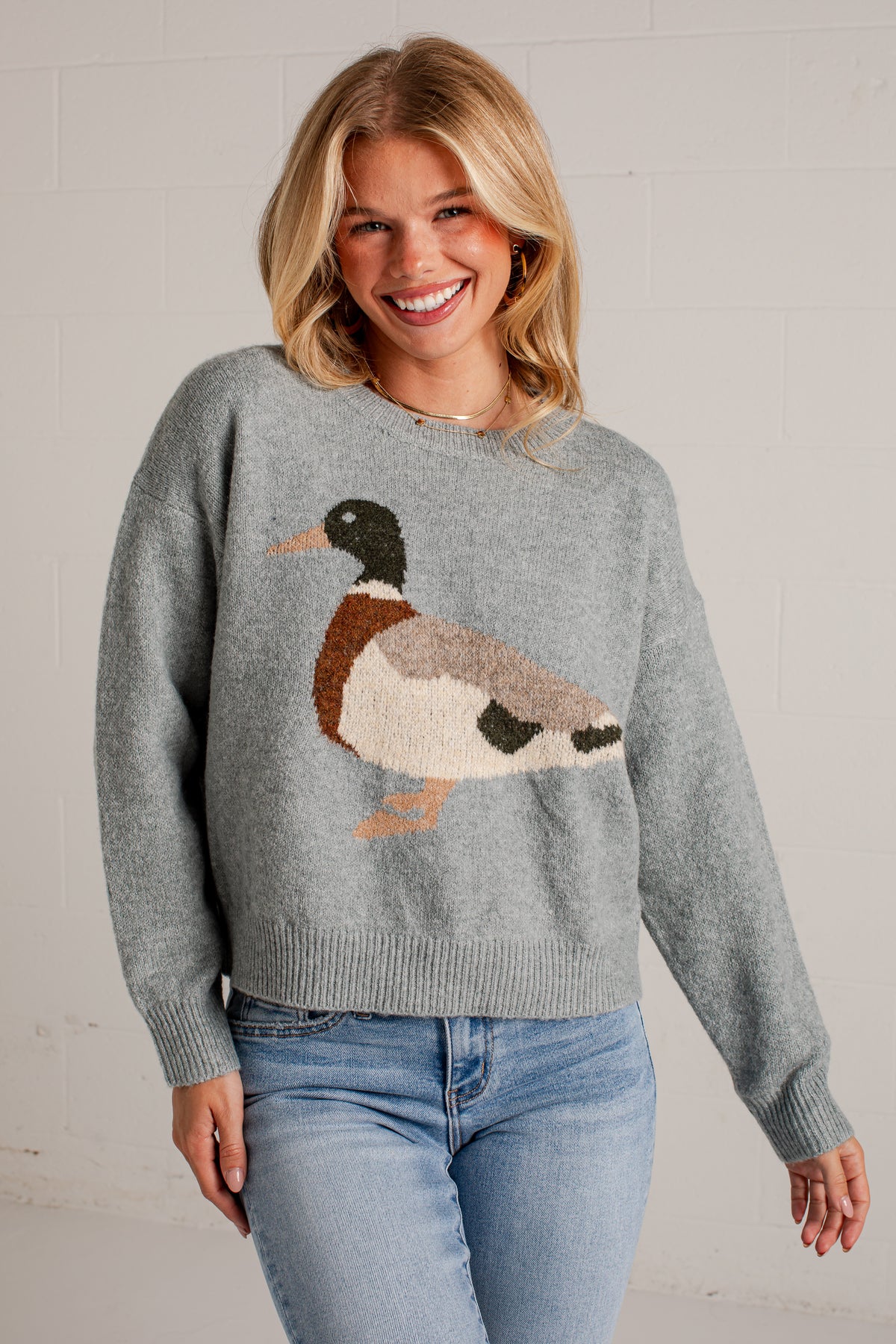 A smiling blonde woman wearing a gray knit sweater with a large duck motif in brown, cream, black, and orange, paired with light blue jeans, standing against a white brick wall.