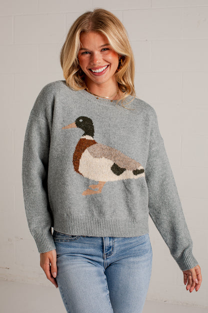 A smiling blonde woman wearing a gray knit sweater with a large duck motif in brown, cream, black, and orange, paired with light blue jeans, standing against a white brick wall.