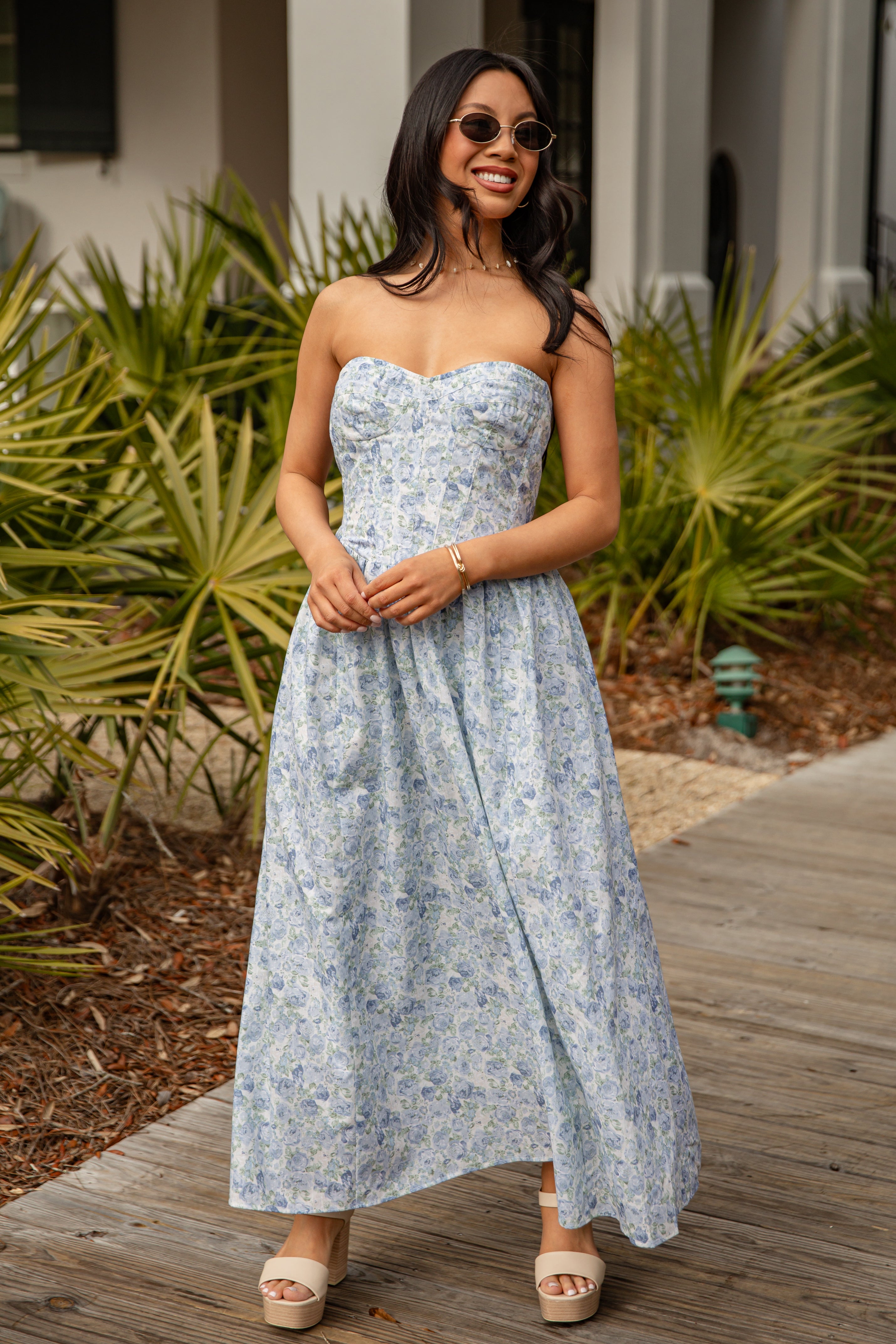 Woman in a strapless floral dress standing outdoors with greenery and a building in the background.