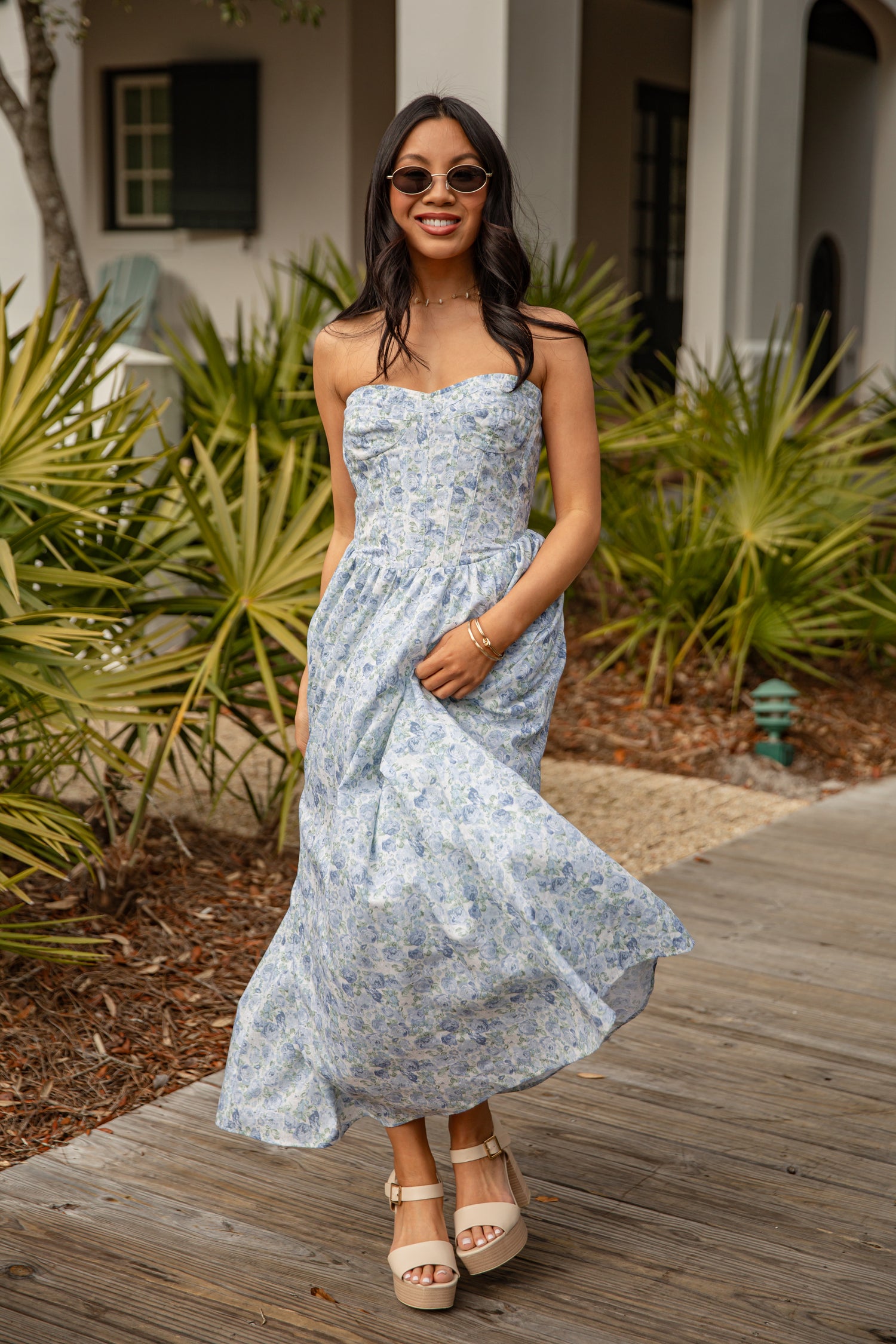 Woman in a light blue floral dress standing outdoors with plants and a building in the background