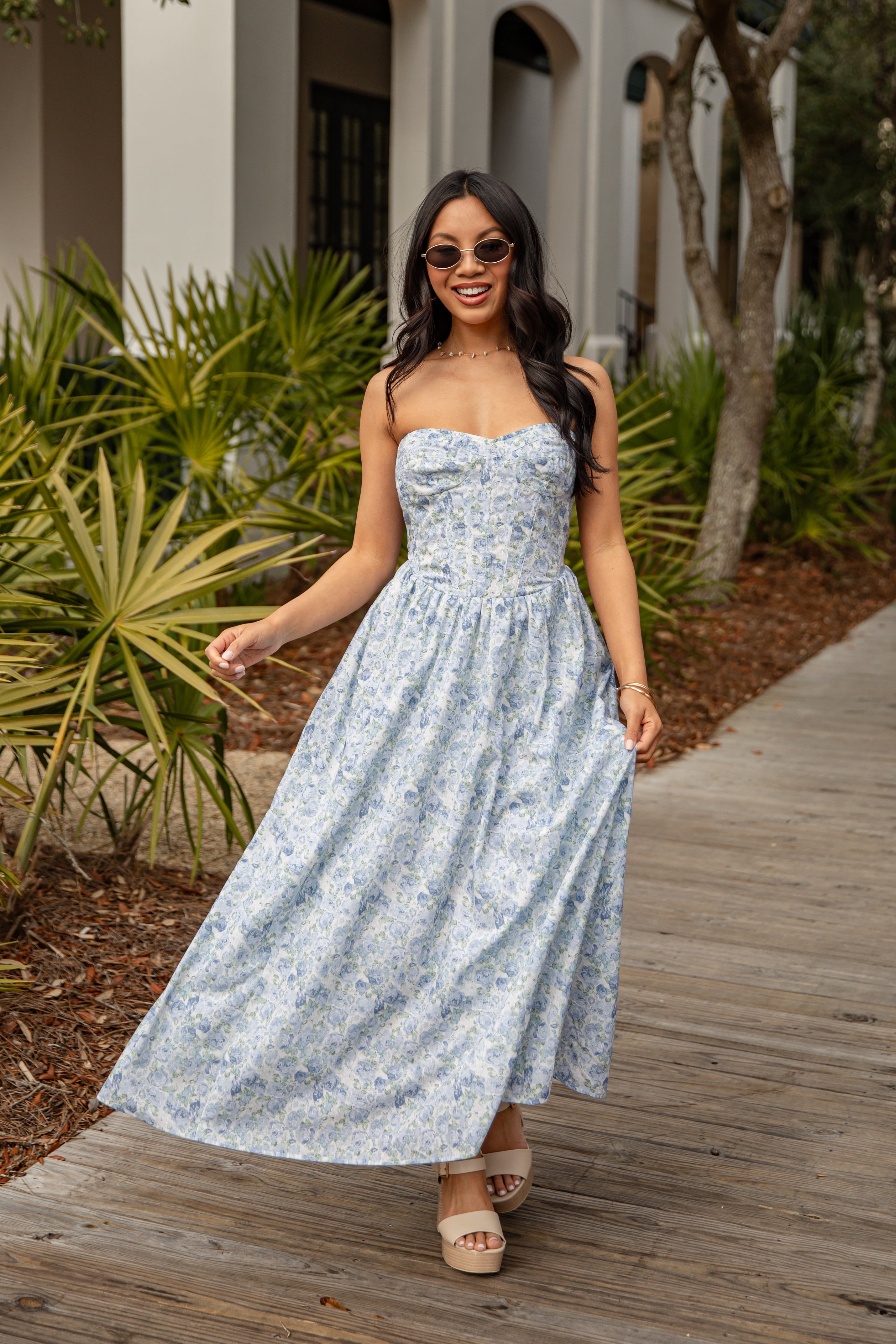 Woman in a floral dress walking on a wooden path with plants and a building in the background