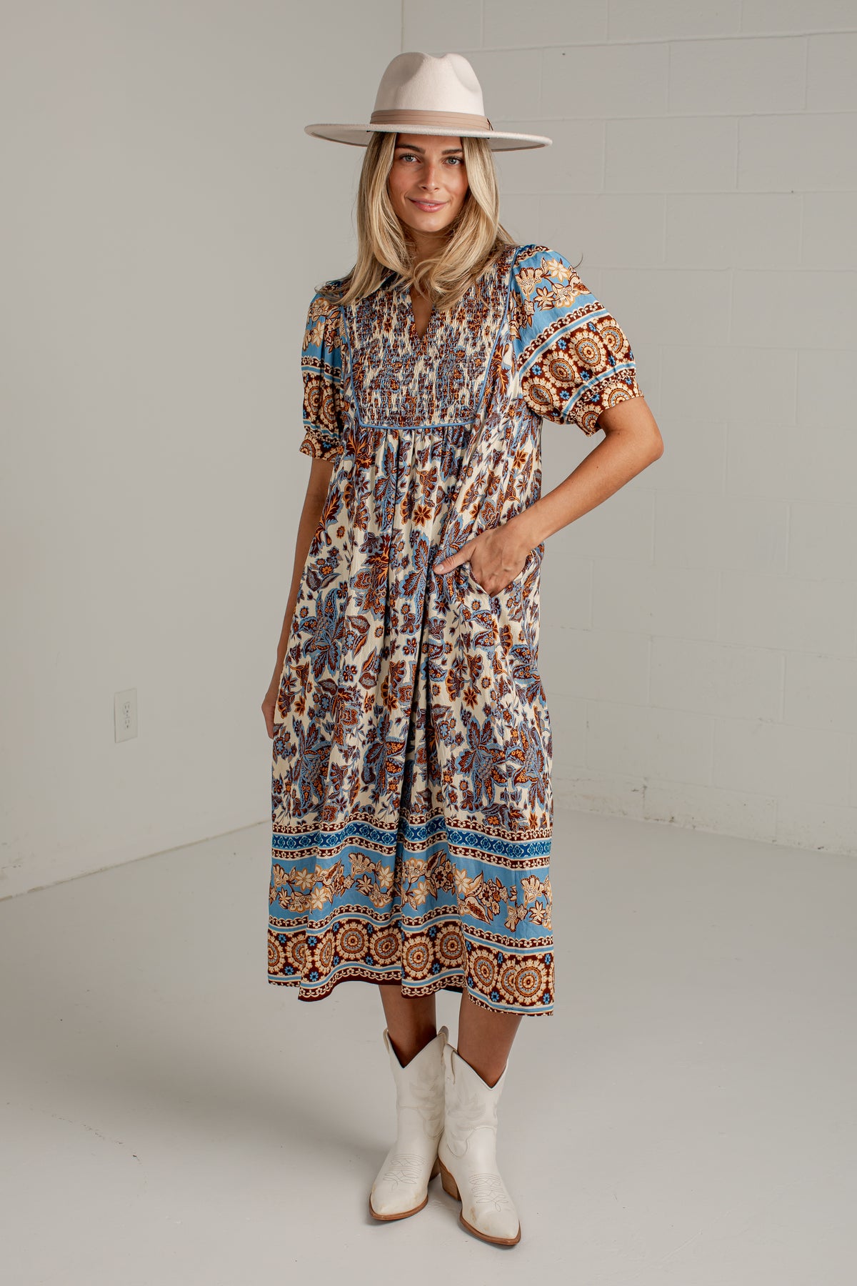 A woman modeling a bohemian-style midi dress with puff sleeves, a gathered waist, and a colorful paisley border. She is wearing a wide-brimmed beige hat and white cowboy boots against a light gray studio background.