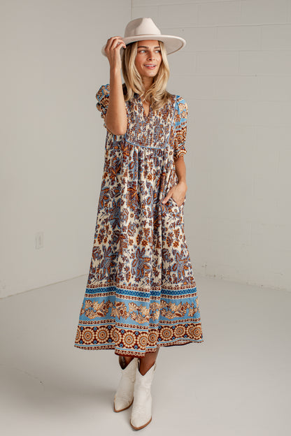 A woman modeling a bohemian-style midi dress with a brown, blue, and beige floral pattern, paired with a beige wide-brim hat and white cowboy boots, standing against a white studio wall.