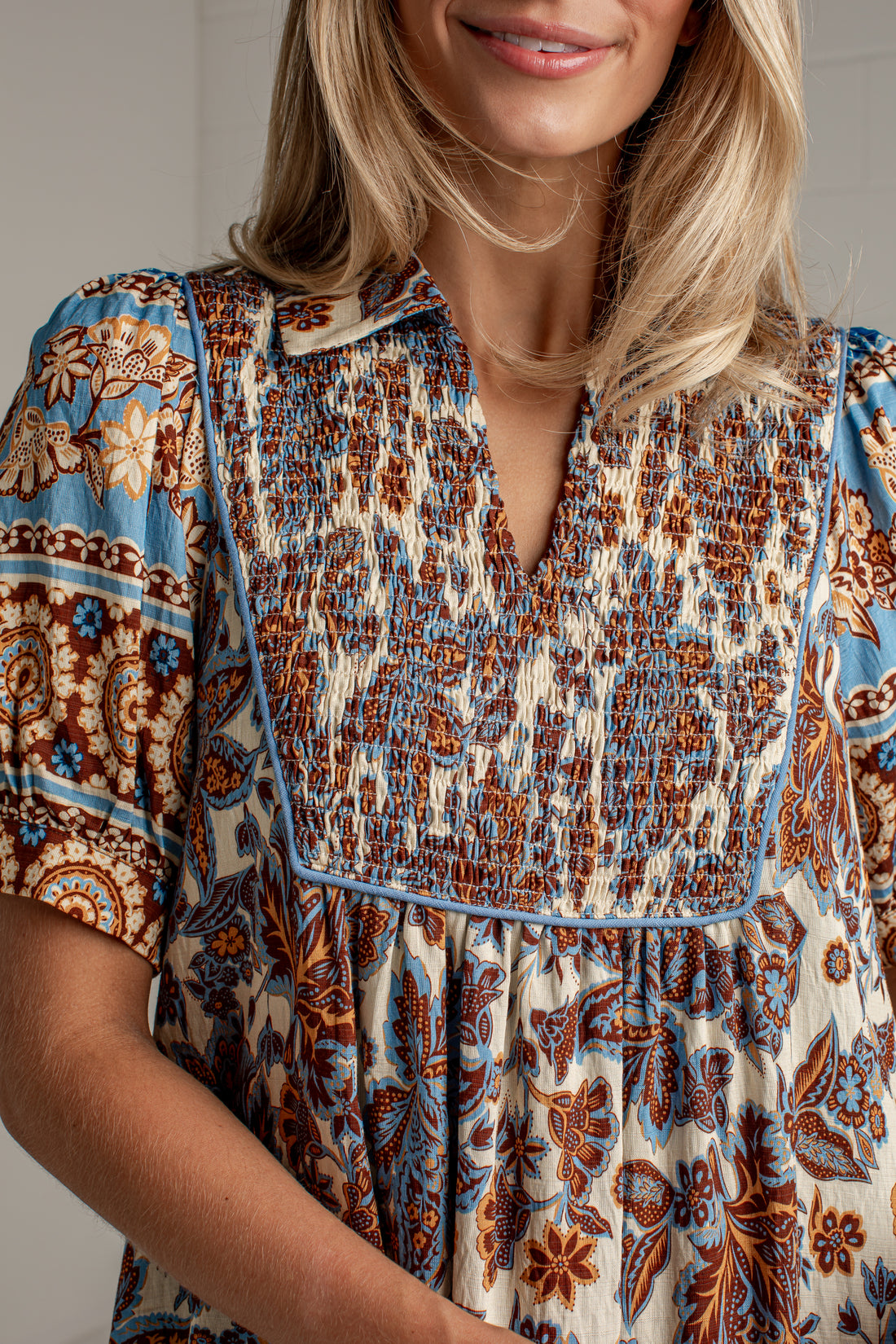 A close-up of a woman wearing a short-sleeve bohemian-style blouse with a smocked chest panel and a blue, brown, and cream floral pattern.