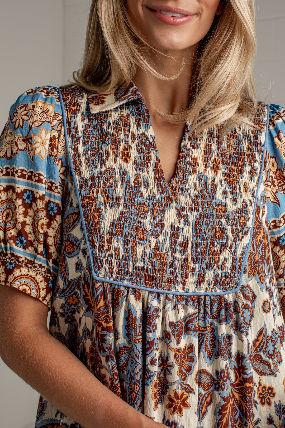 A close-up of a woman wearing a short-sleeve bohemian-style blouse with a smocked chest panel and a blue, brown, and cream floral pattern.