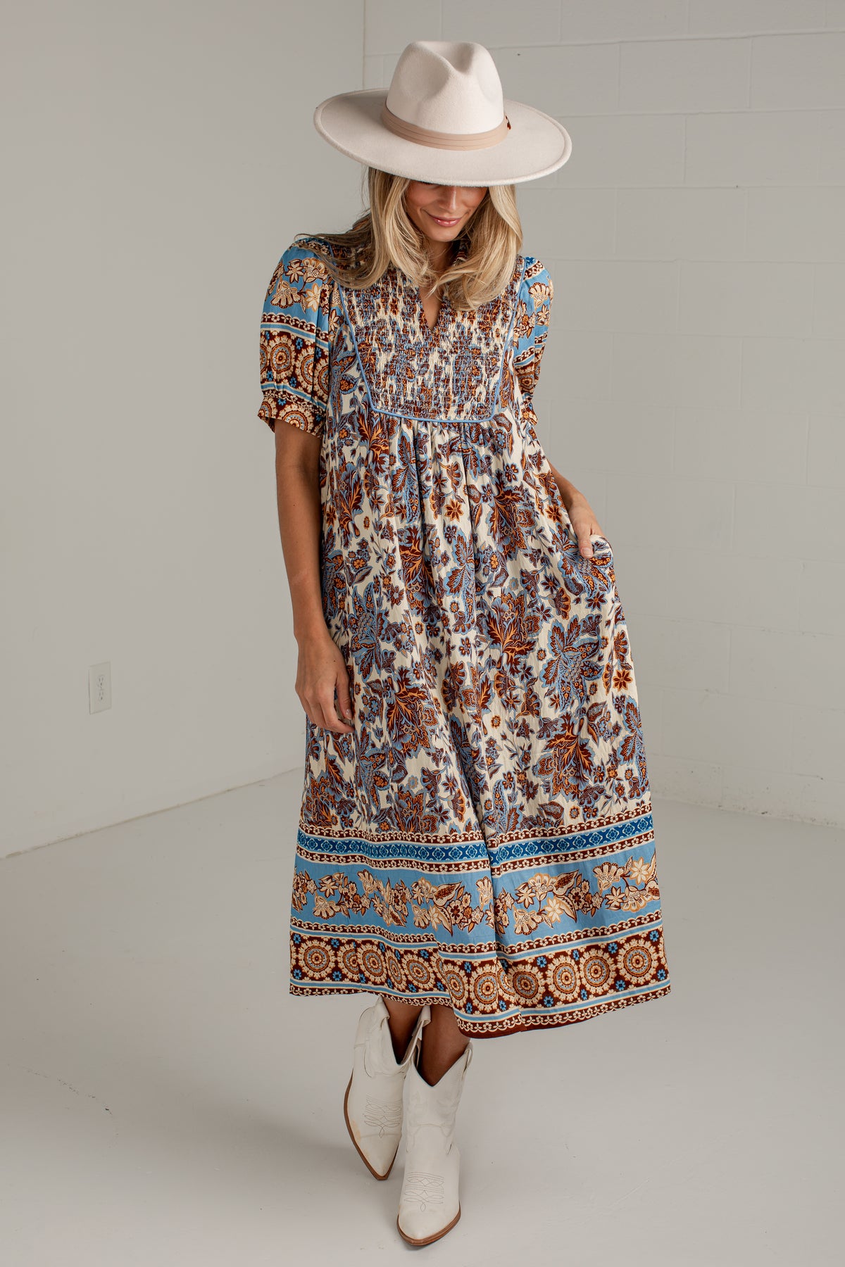 A woman modeling a bohemian-style midi dress with a smocked bodice and a colorful paisley border, paired with a wide-brimmed beige hat and white cowboy boots.