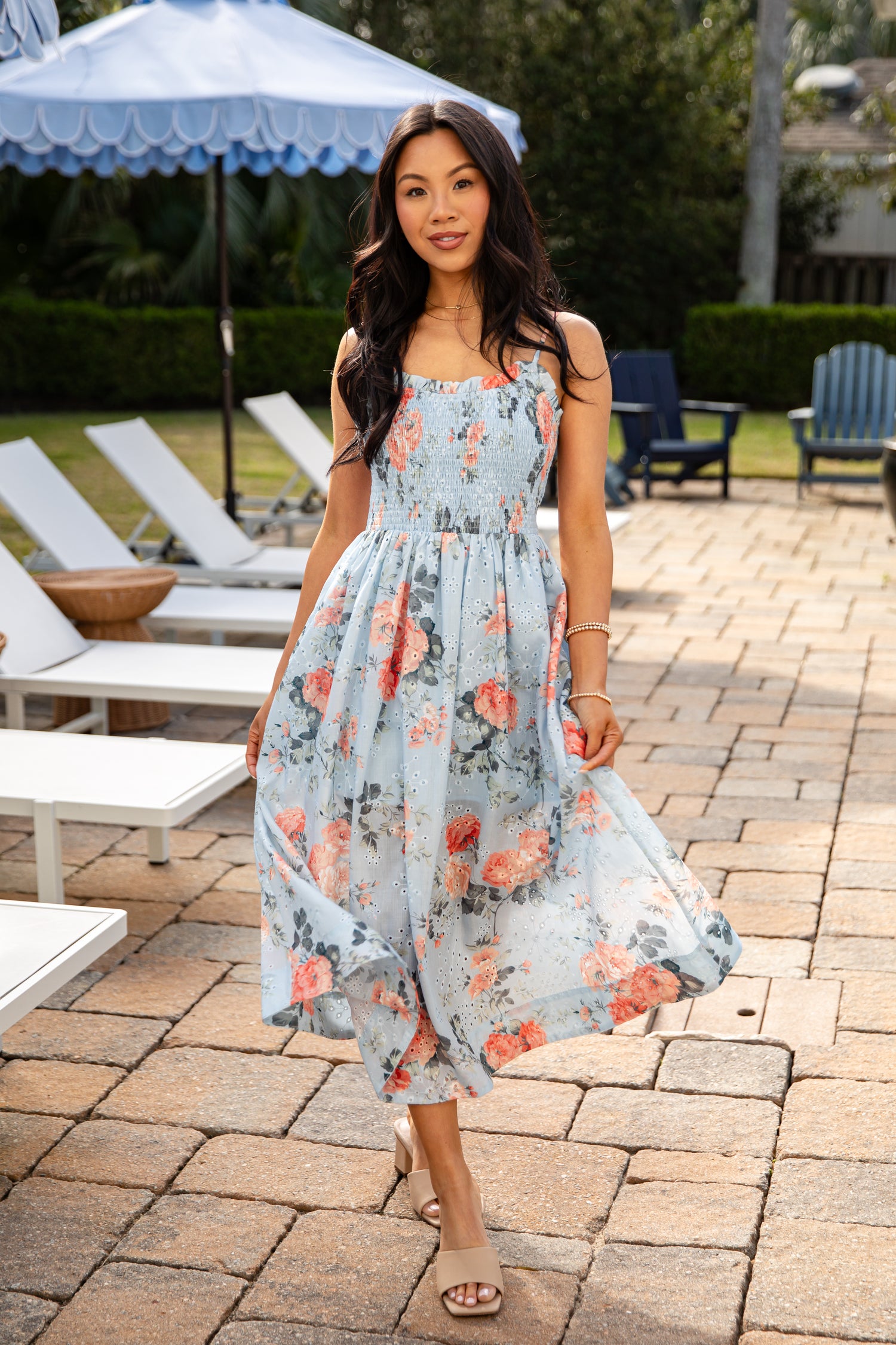 Woman in a floral dress standing outdoors near lounge chairs and an umbrella.
