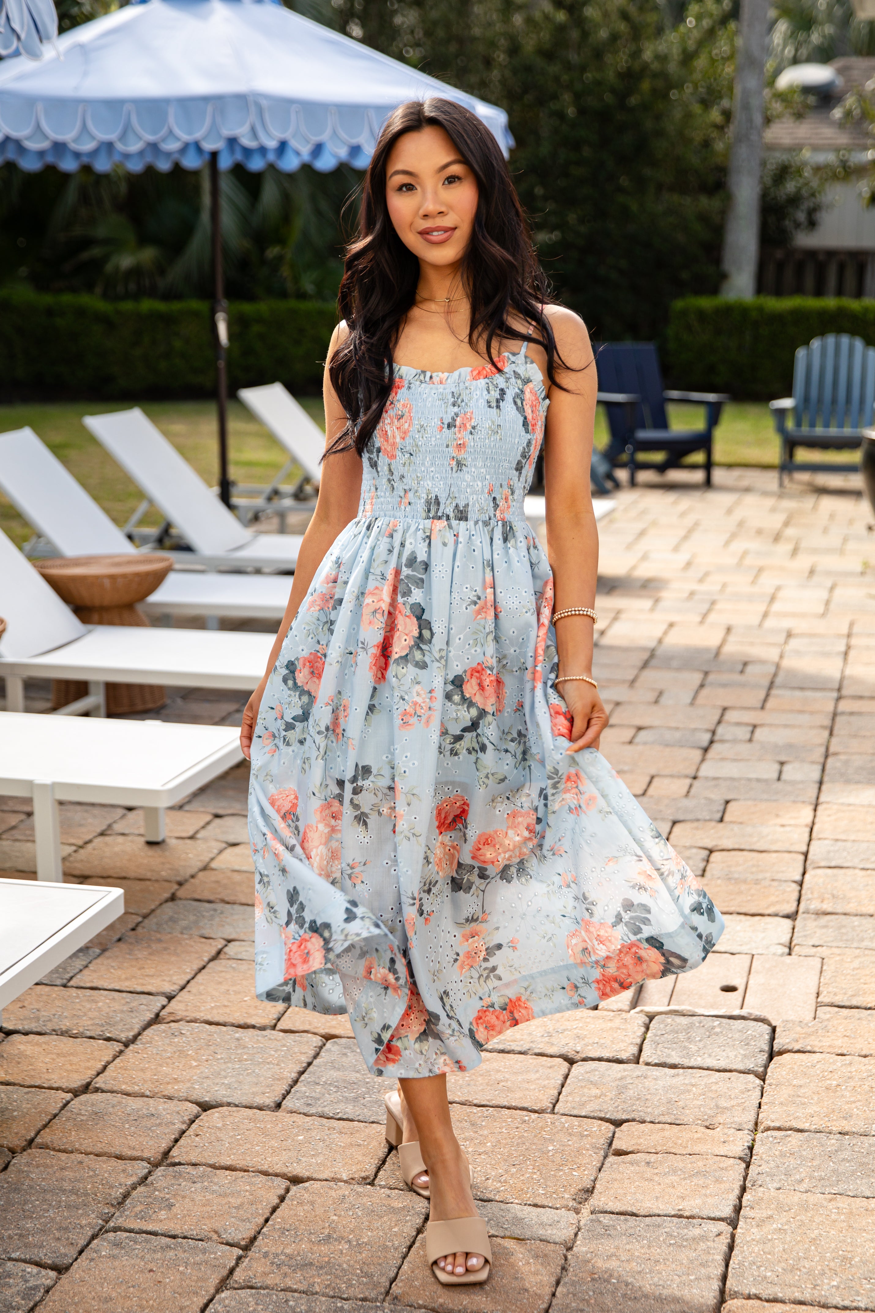 Woman in a floral dress standing outdoors near lounge chairs and an umbrella.