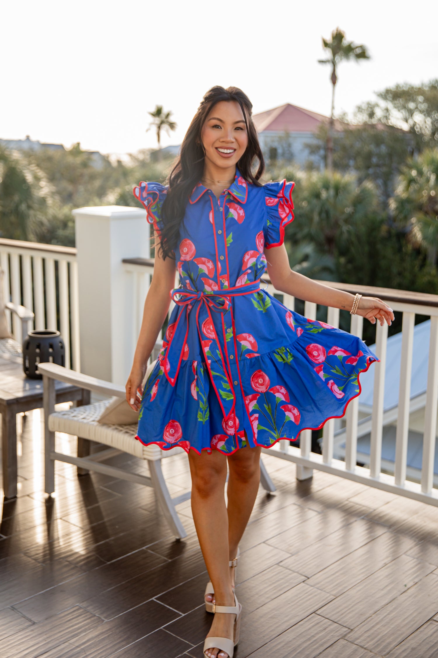 Woman in a blue floral dress standing on a balcony with greenery and buildings in the background.