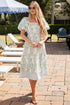 Woman in a floral dress standing on a patio with outdoor furniture and greenery.