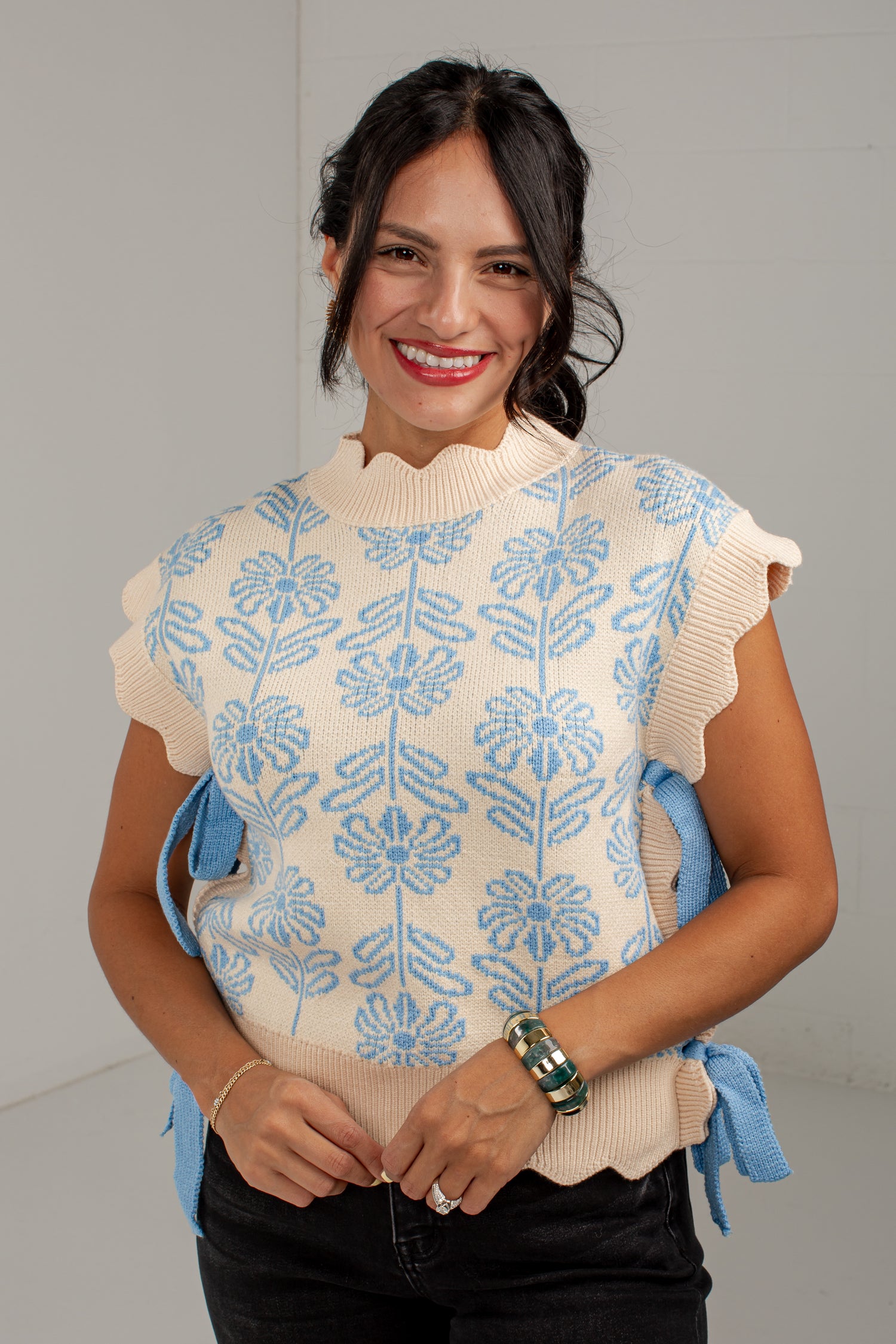 A smiling woman wearing a cream knit sweater vest with a light blue floral pattern, scalloped edges, and blue side ties, paired with black pants.