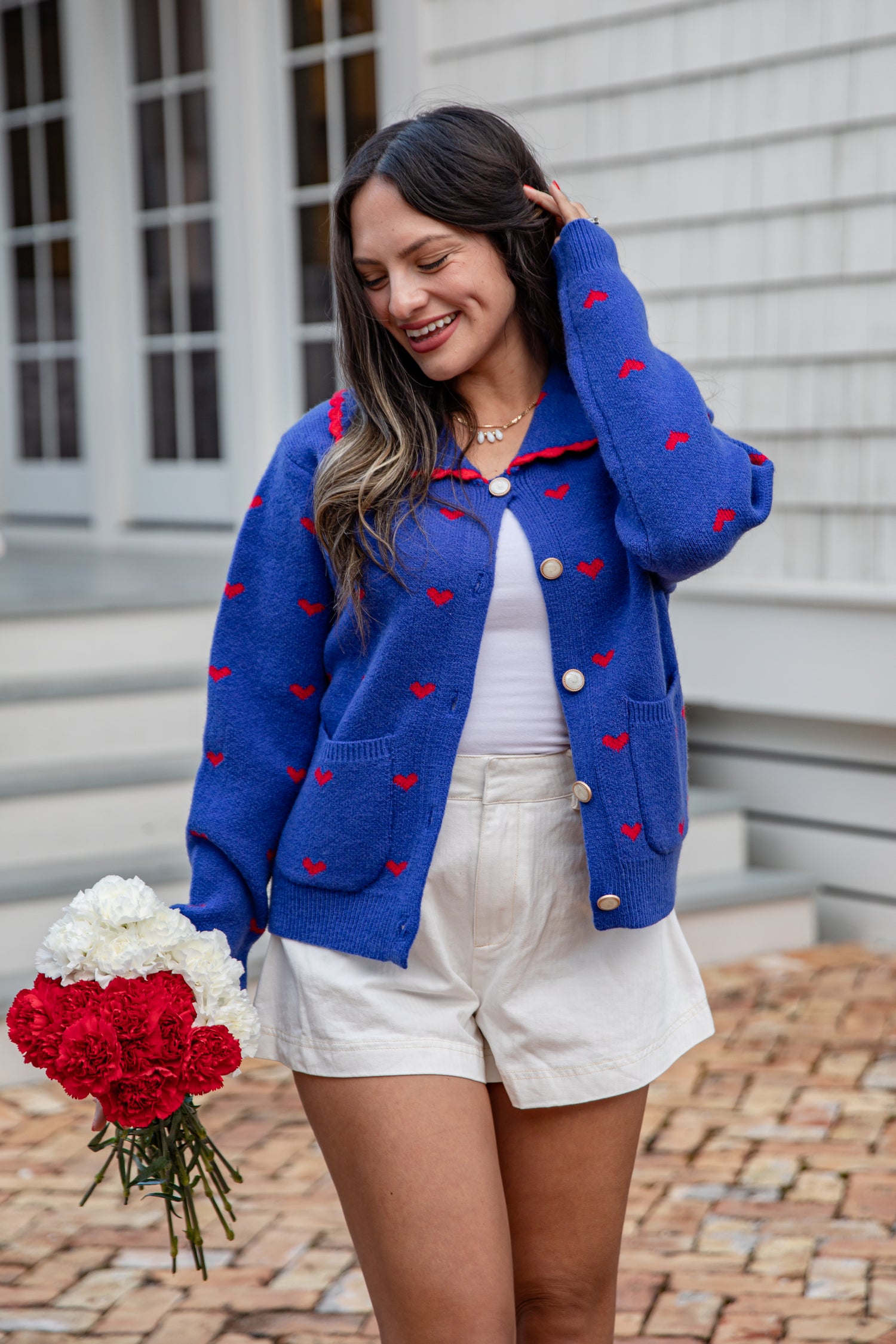 Woman wearing a blue cardigan with red heart patterns, white shorts, and holding flowers.