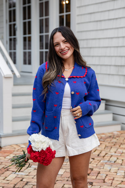 Woman wearing a blue cardigan with red accents, holding flowers, standing outside a house.