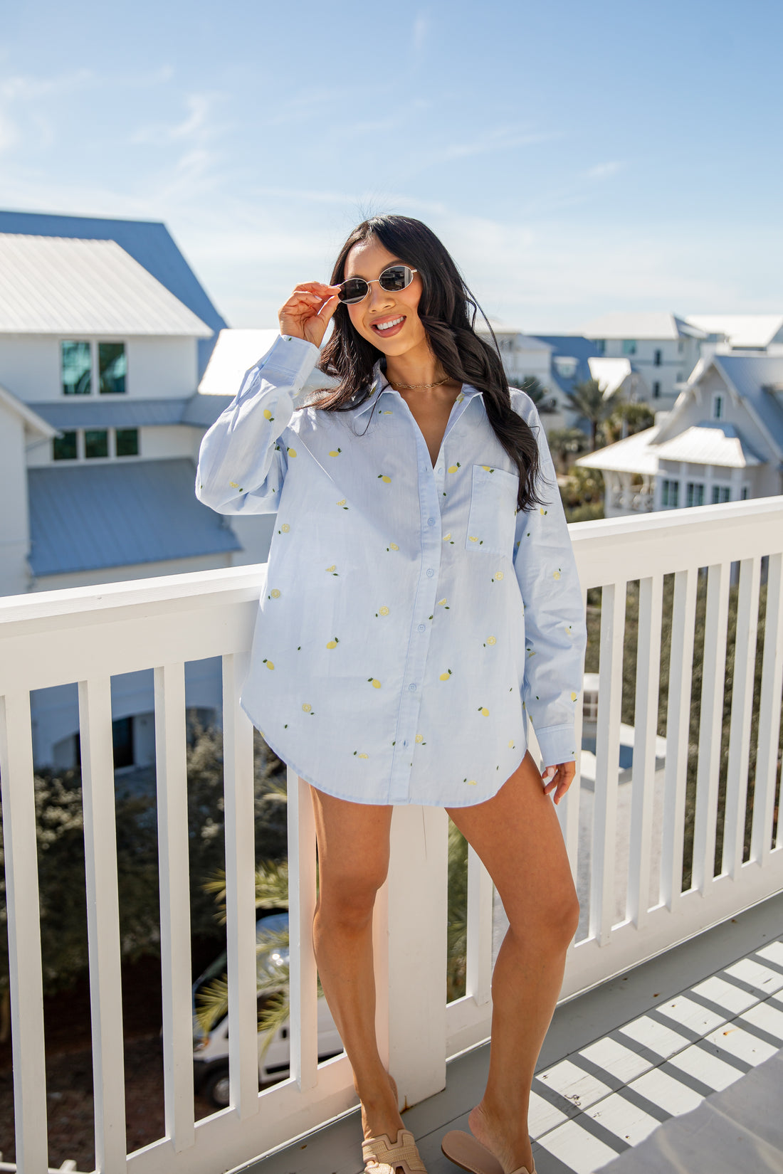 Woman in a light blue shirt with a pattern, standing on a balcony with a coastal view.