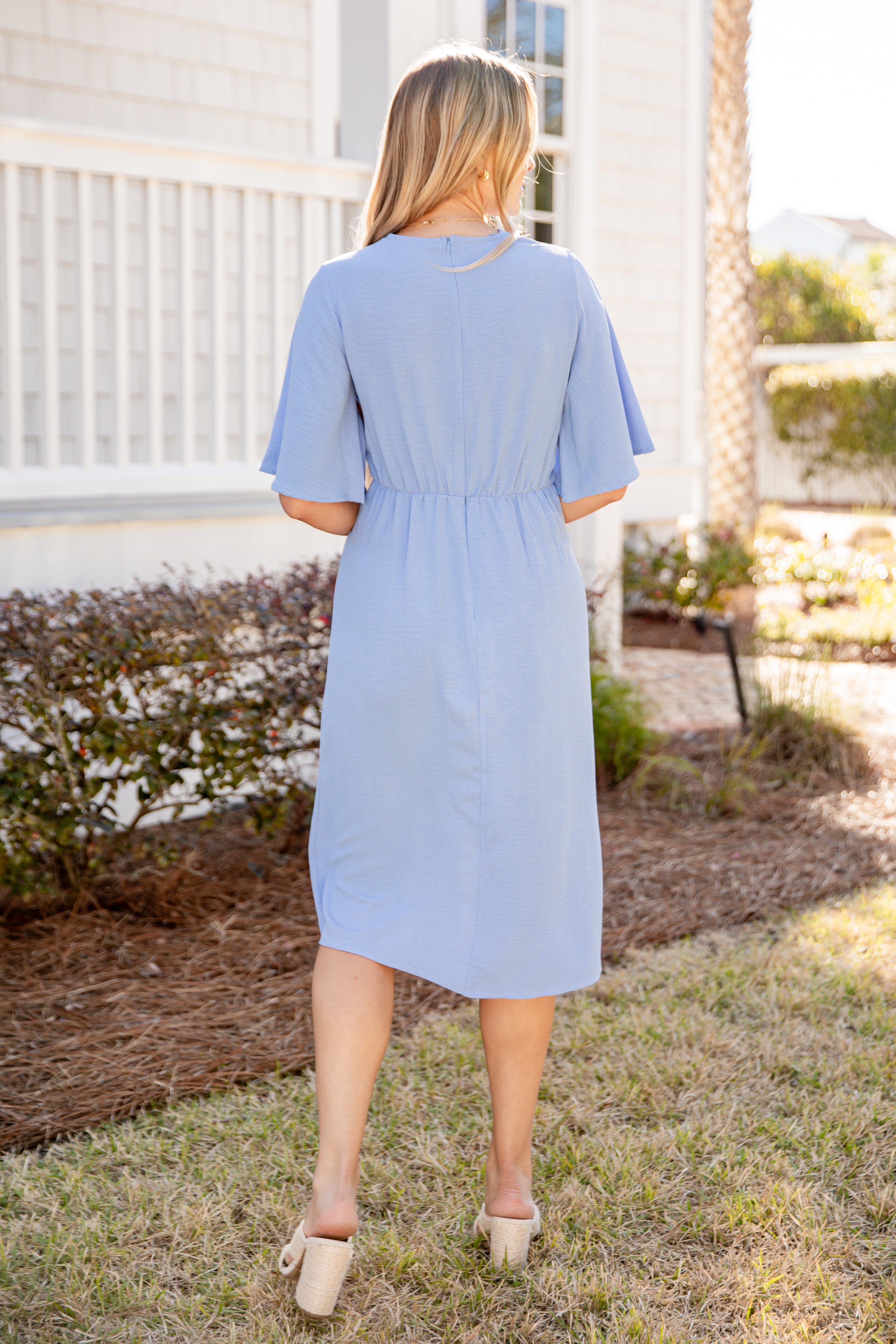 Woman wearing a light blue dress standing outdoors with a white building and palm tree in the background.