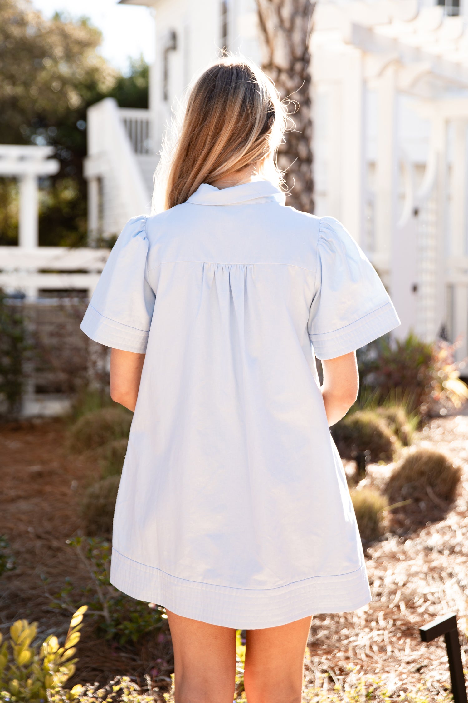Woman wearing a light blue dress standing outdoors with greenery and a white building in the background