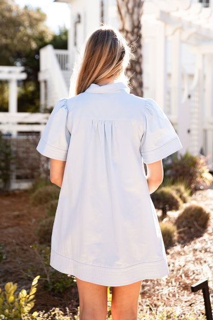 Woman wearing a light blue dress standing outdoors with greenery and a white building in the background