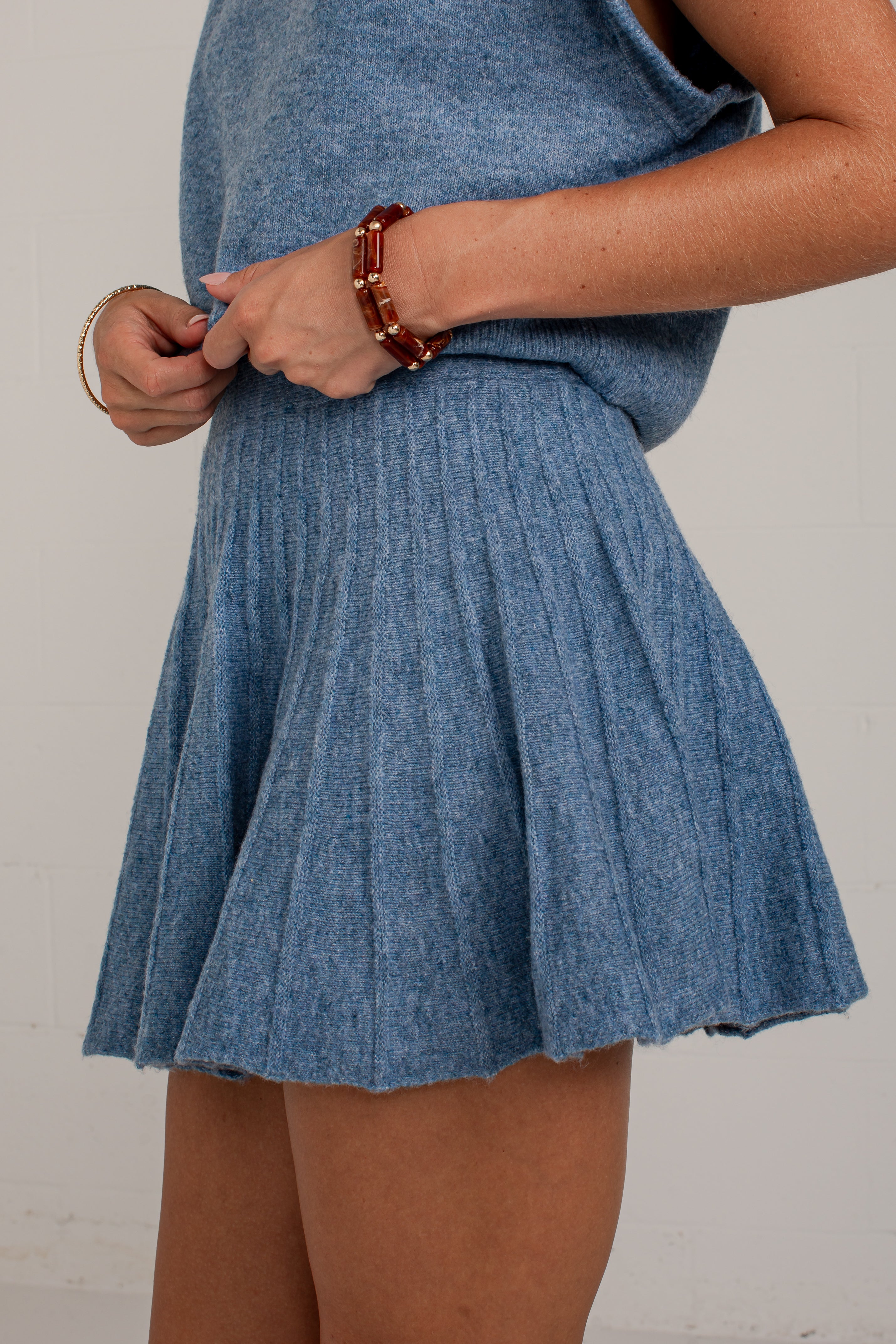 A person wearing a heathered blue knit sleeveless top and matching pleated mini skirt, shown from the chest down against a plain light wall.