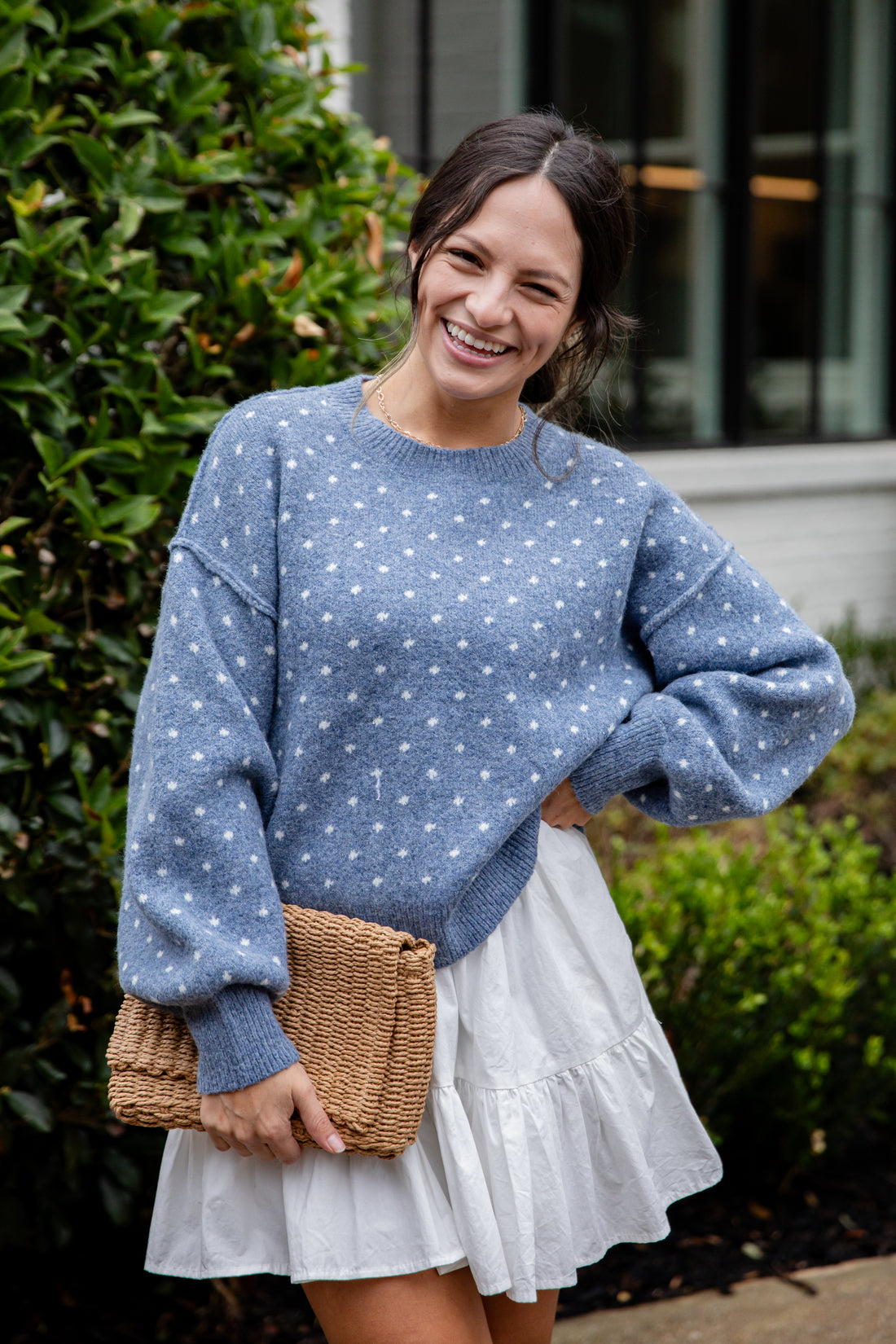 Woman wearing a blue polka dot sweater and white skirt, holding a woven bag outdoors.