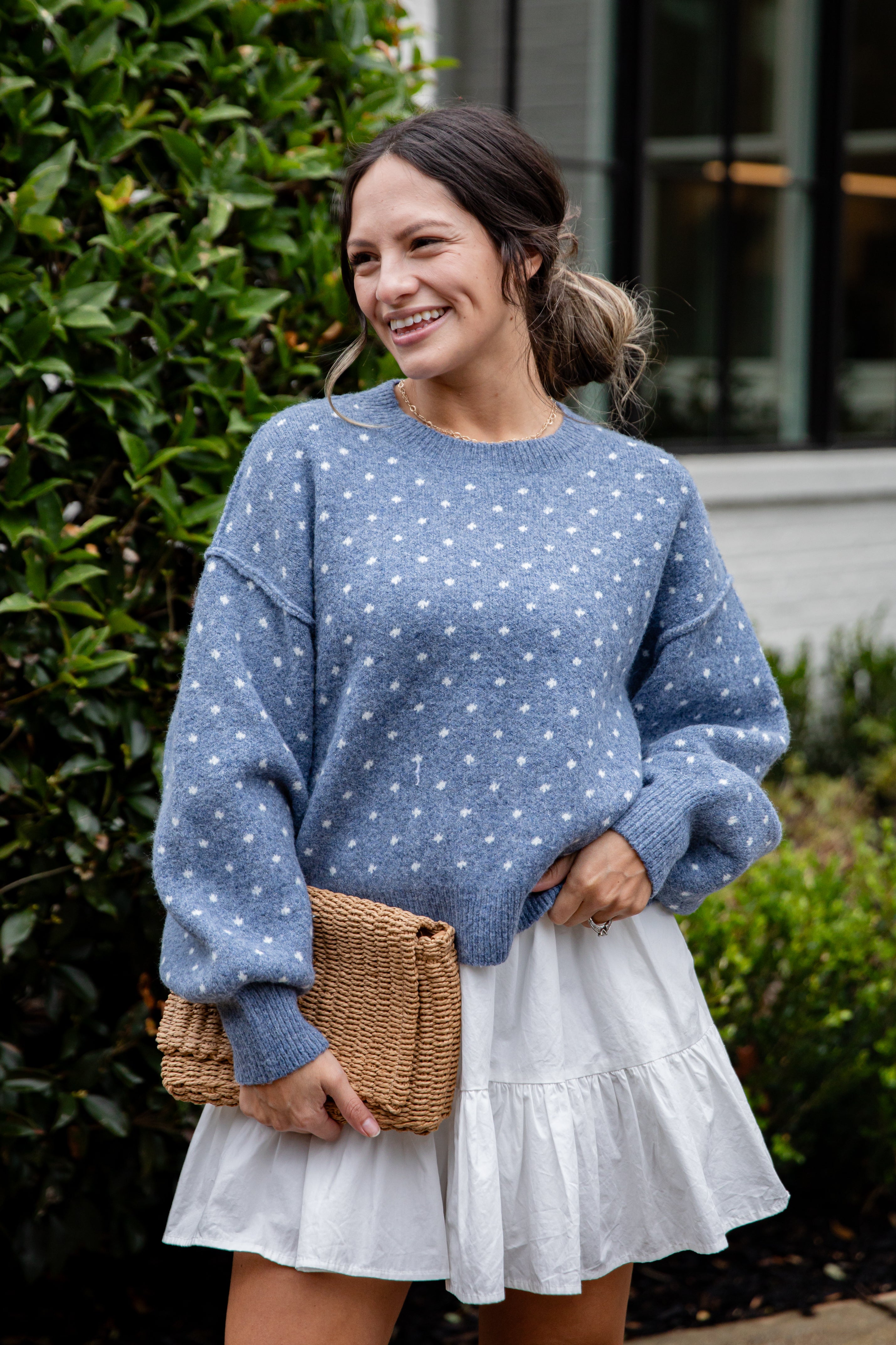 Woman wearing a blue polka dot sweater and white skirt holding a woven bag outdoors.