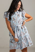 A woman wearing a light blue short-sleeve button-up dress with a brown and white floral print, ruffled sleeves, and a matching fabric tie belt at the waist, standing against a light gray wall.