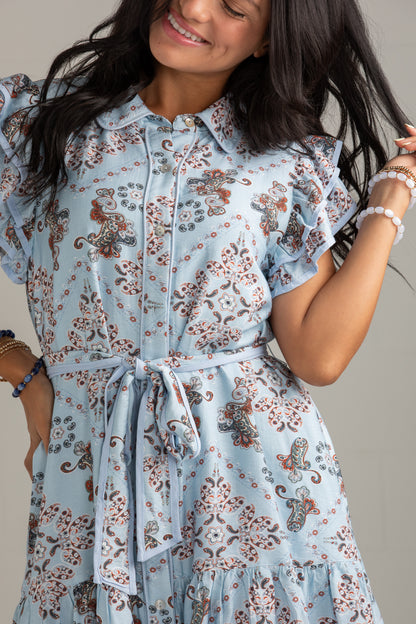 A woman wearing a light blue short-sleeve button-up dress with a brown and beige floral pattern, featuring ruffled sleeves and a matching fabric tie belt at the waist.
