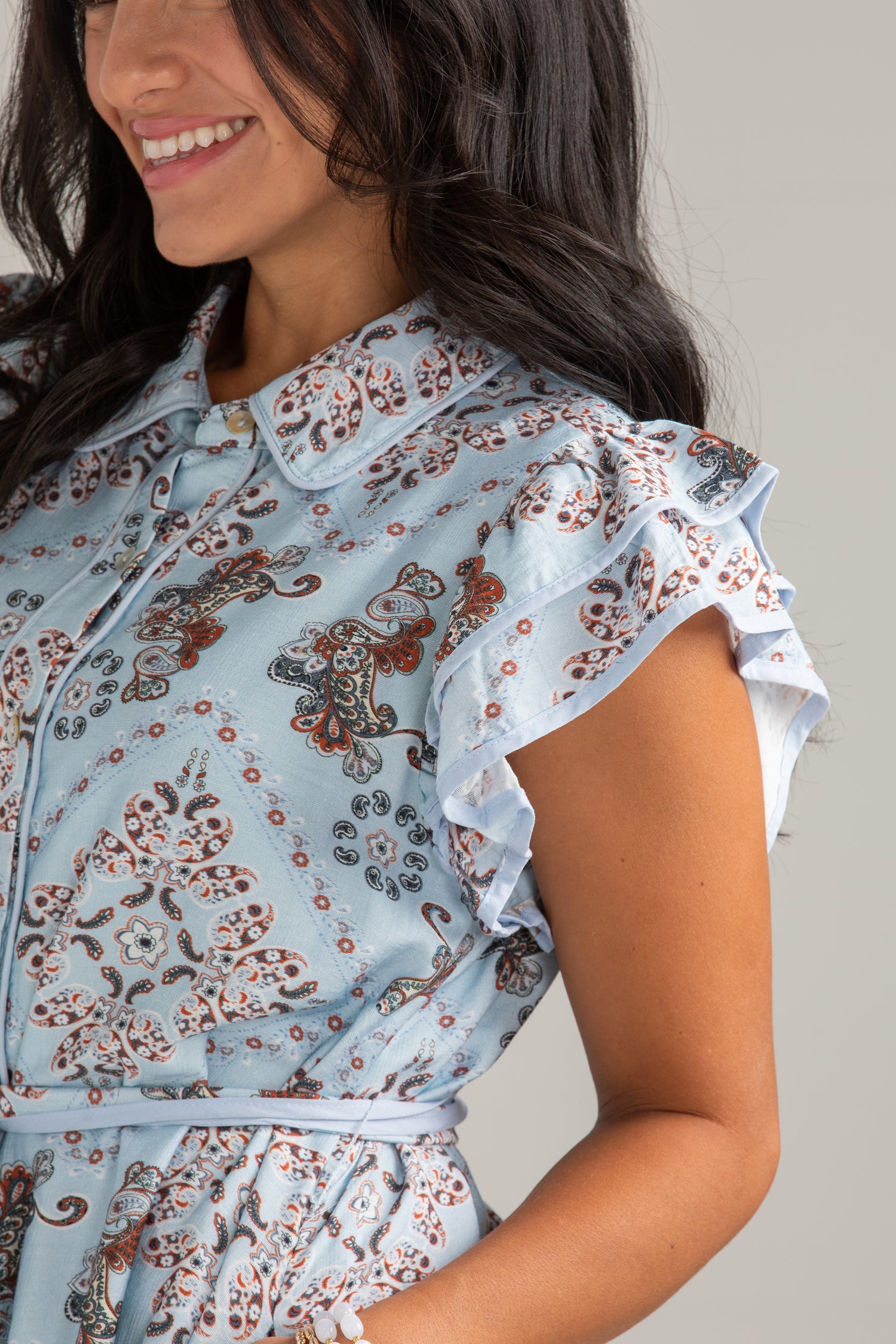 A close-up of a woman wearing a light blue ruffled blouse with a brown and gray paisley pattern, featuring a collar and buttoned front.