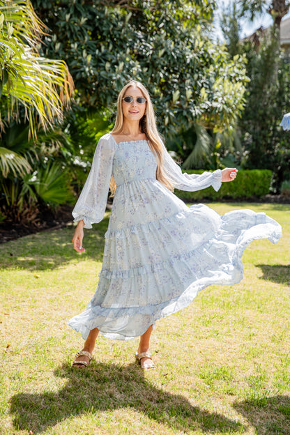 Woman in a flowing light blue dress standing on grass with trees in the background
