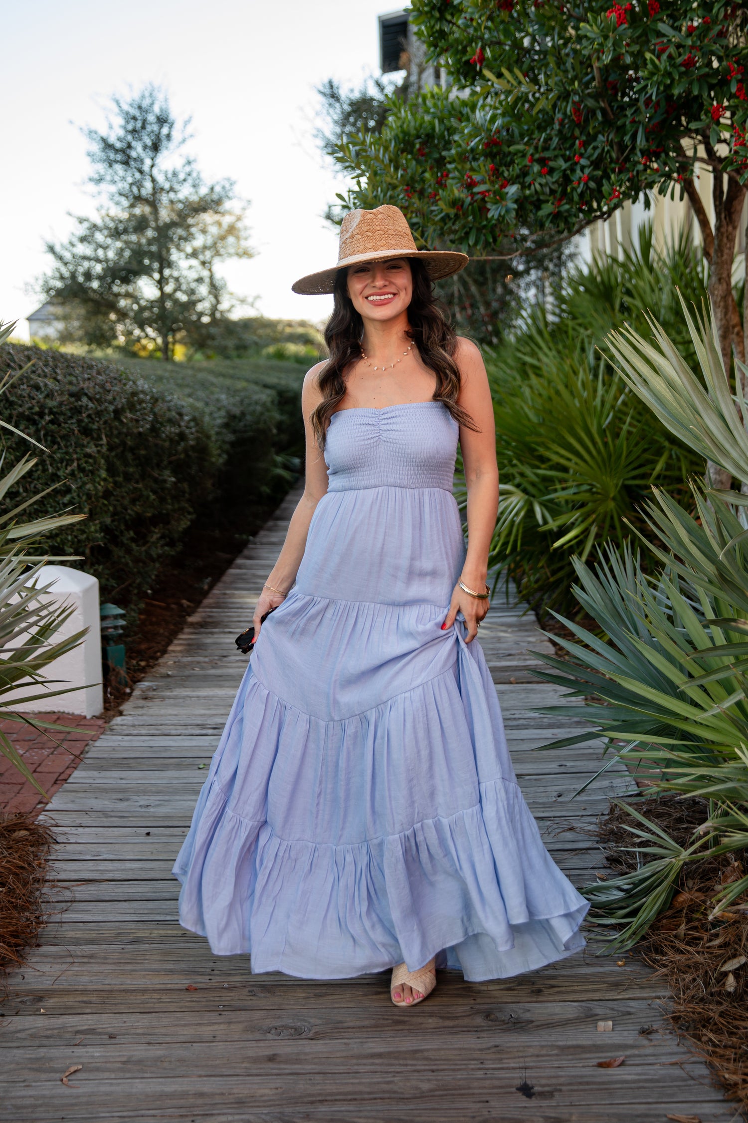 Woman in a light blue dress and hat standing on a wooden path with greenery around.