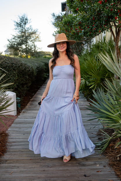 Woman in a light blue dress and hat standing on a wooden path with greenery around.