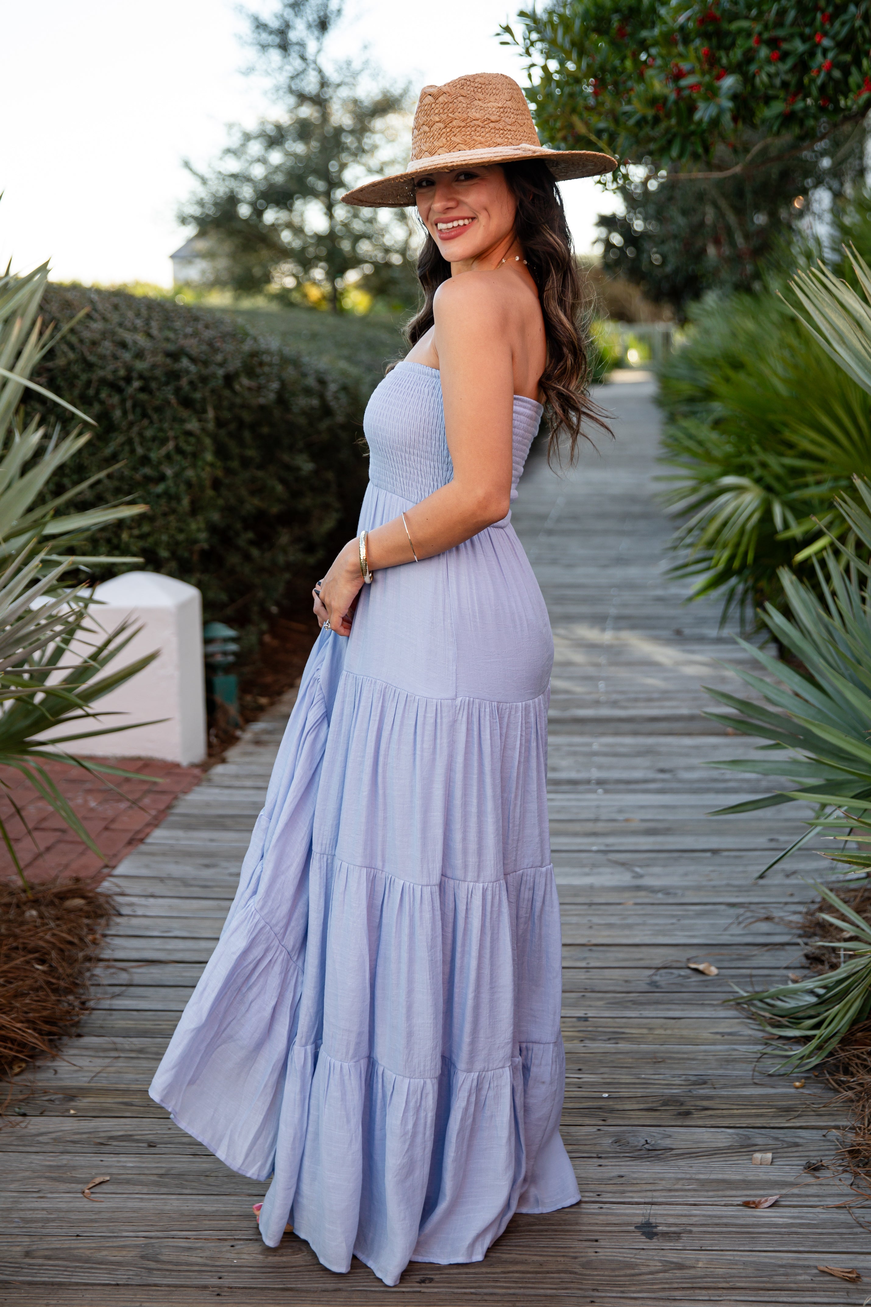 Woman in a light blue dress and straw hat standing on a wooden path with greenery around.