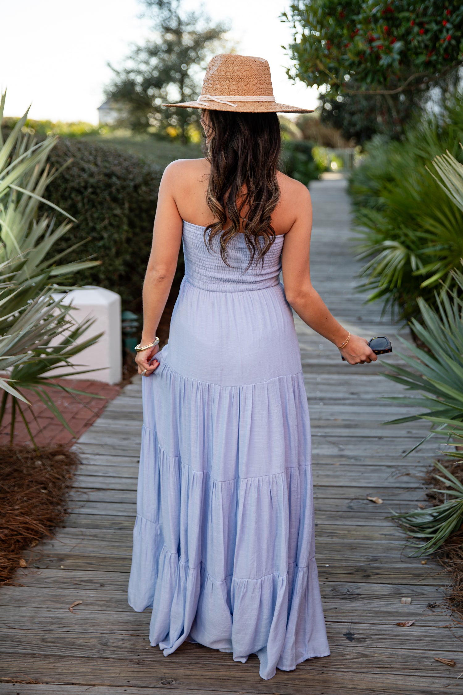 Woman in a long light blue dress and straw hat walking on a wooden path in a garden.