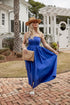 Woman in a blue dress and hat standing on a brick patio with a white house in the background