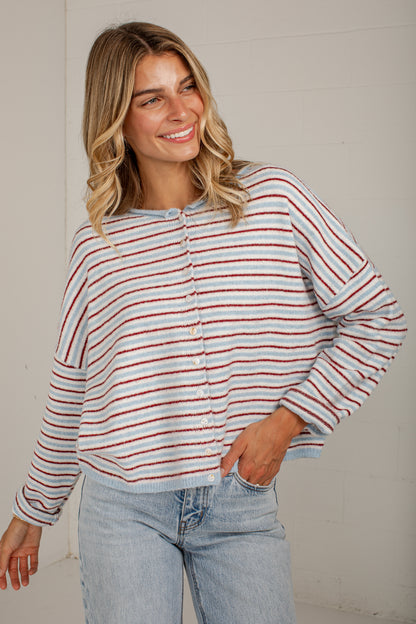 A woman wearing a light blue knit cardigan with thin red and white horizontal stripes, paired with light-wash blue jeans, standing against a white paneled wall.