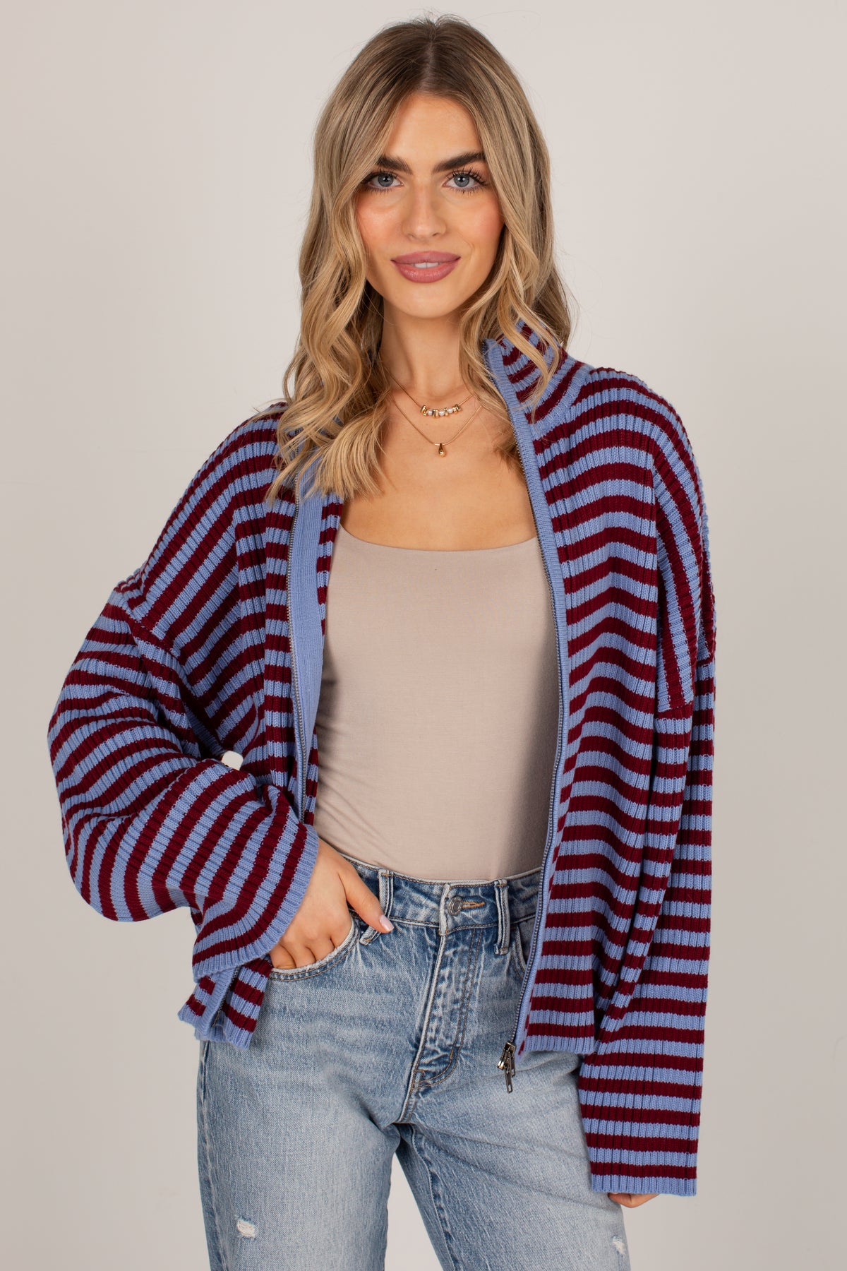 A woman modeling a striped knit zip-up cardigan with blue and burgundy horizontal stripes, worn open over a beige tank top and light-wash blue jeans against a plain light gray studio background.