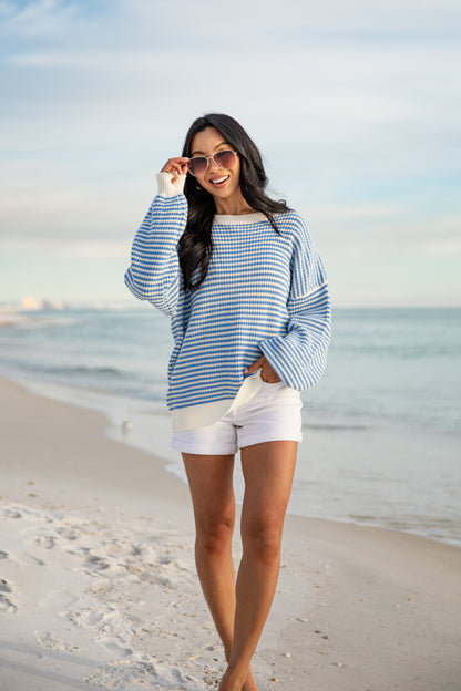 Woman in a blue and white striped sweater and white shorts standing on a beach.