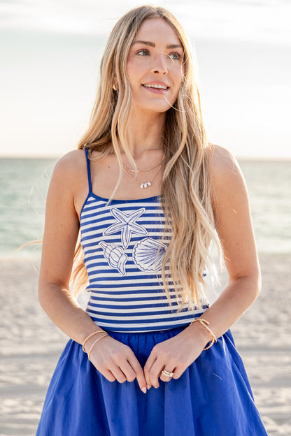 Woman wearing a blue and white striped tank top with shell designs on a beach.