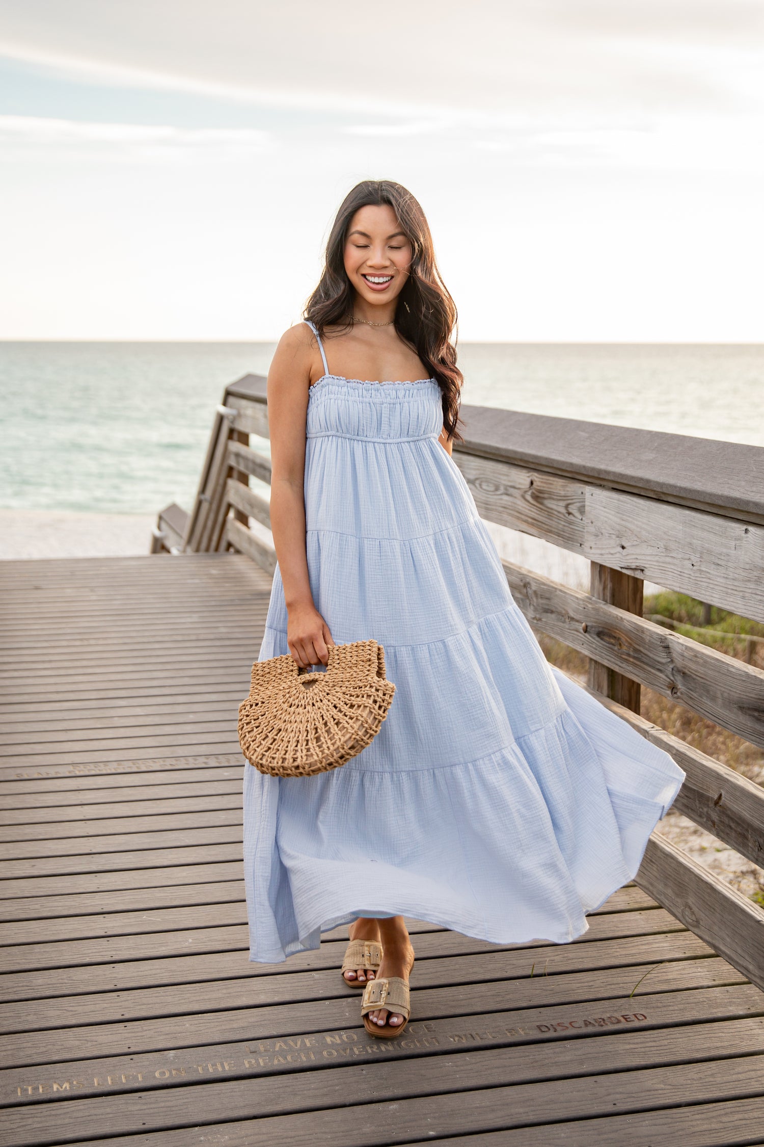 Woman in a light blue dress holding a woven handbag on a wooden deck by the water.