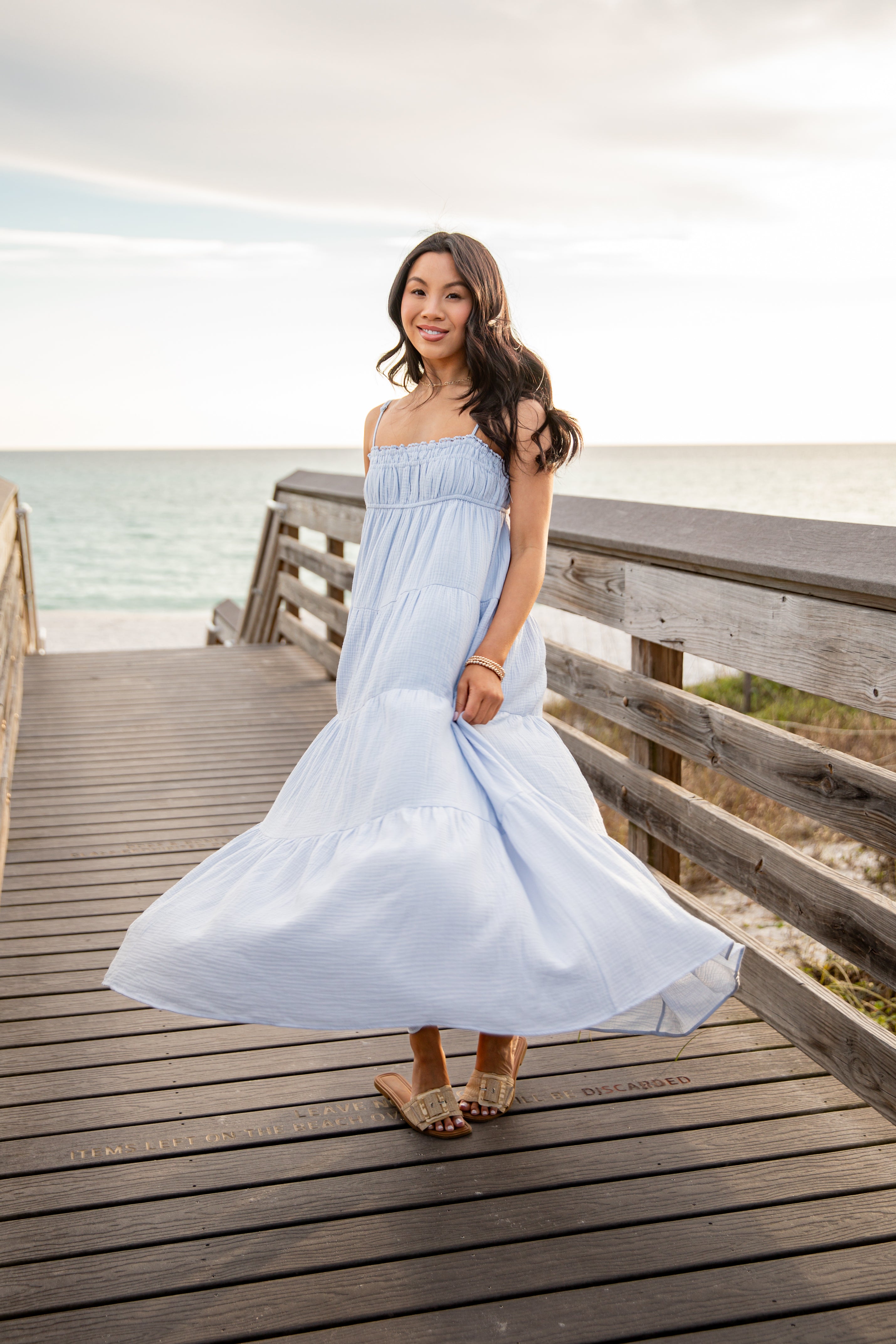 Woman in a light blue dress standing on a wooden boardwalk by the beach.