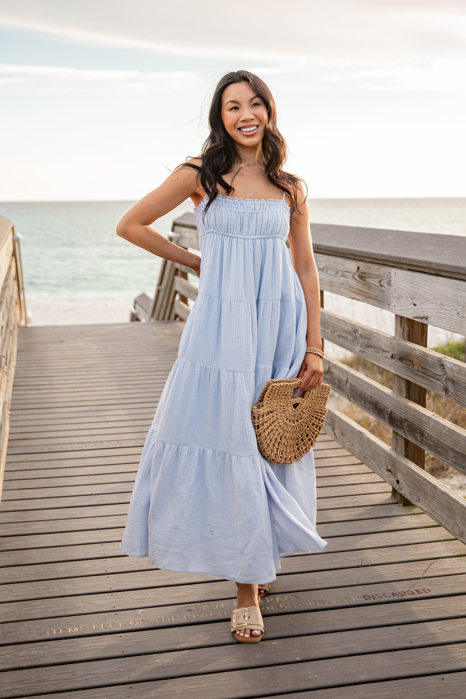 Woman in a light blue dress standing on a wooden boardwalk by the beach