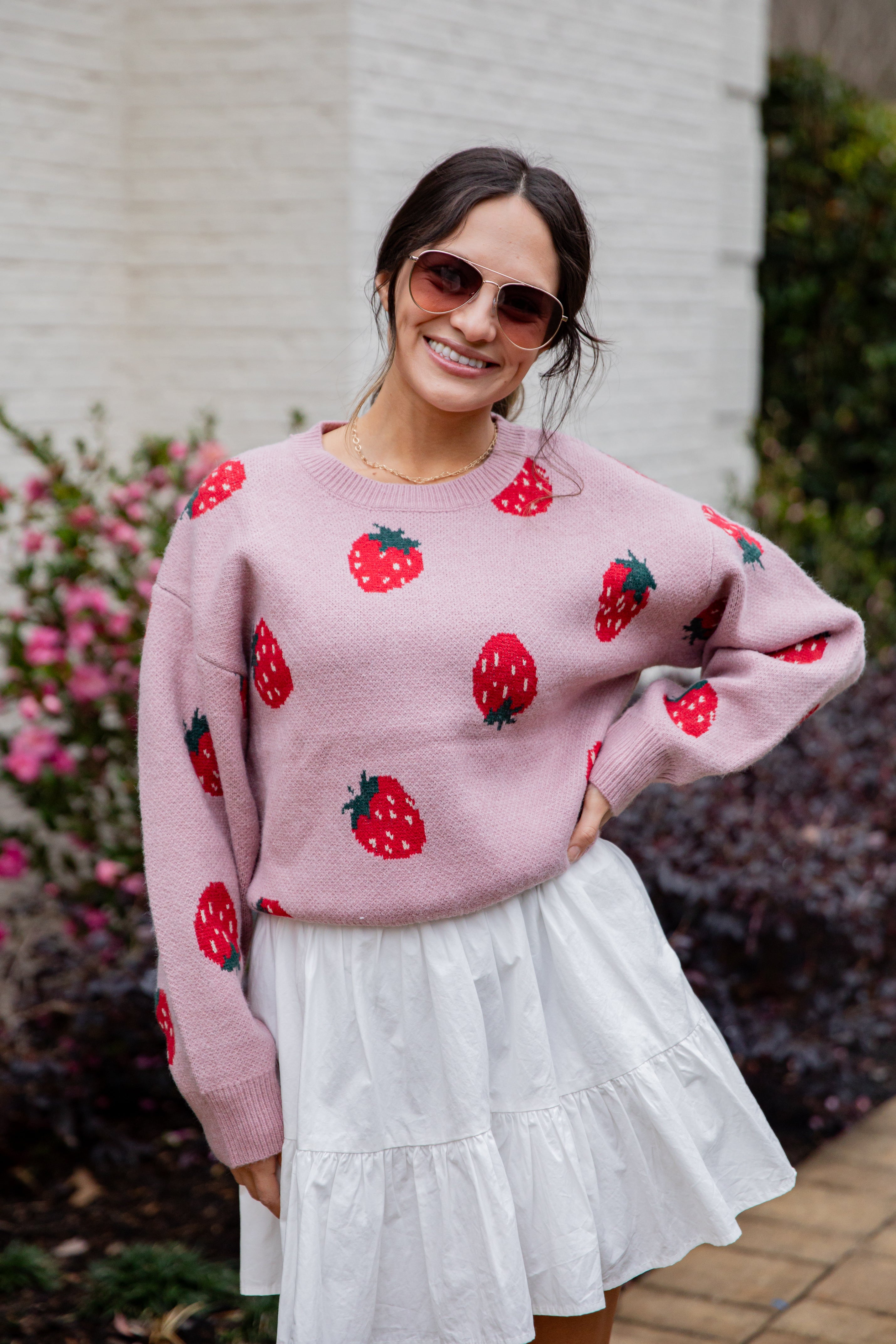 Woman wearing a pink sweater with strawberry patterns and a white skirt outdoors.