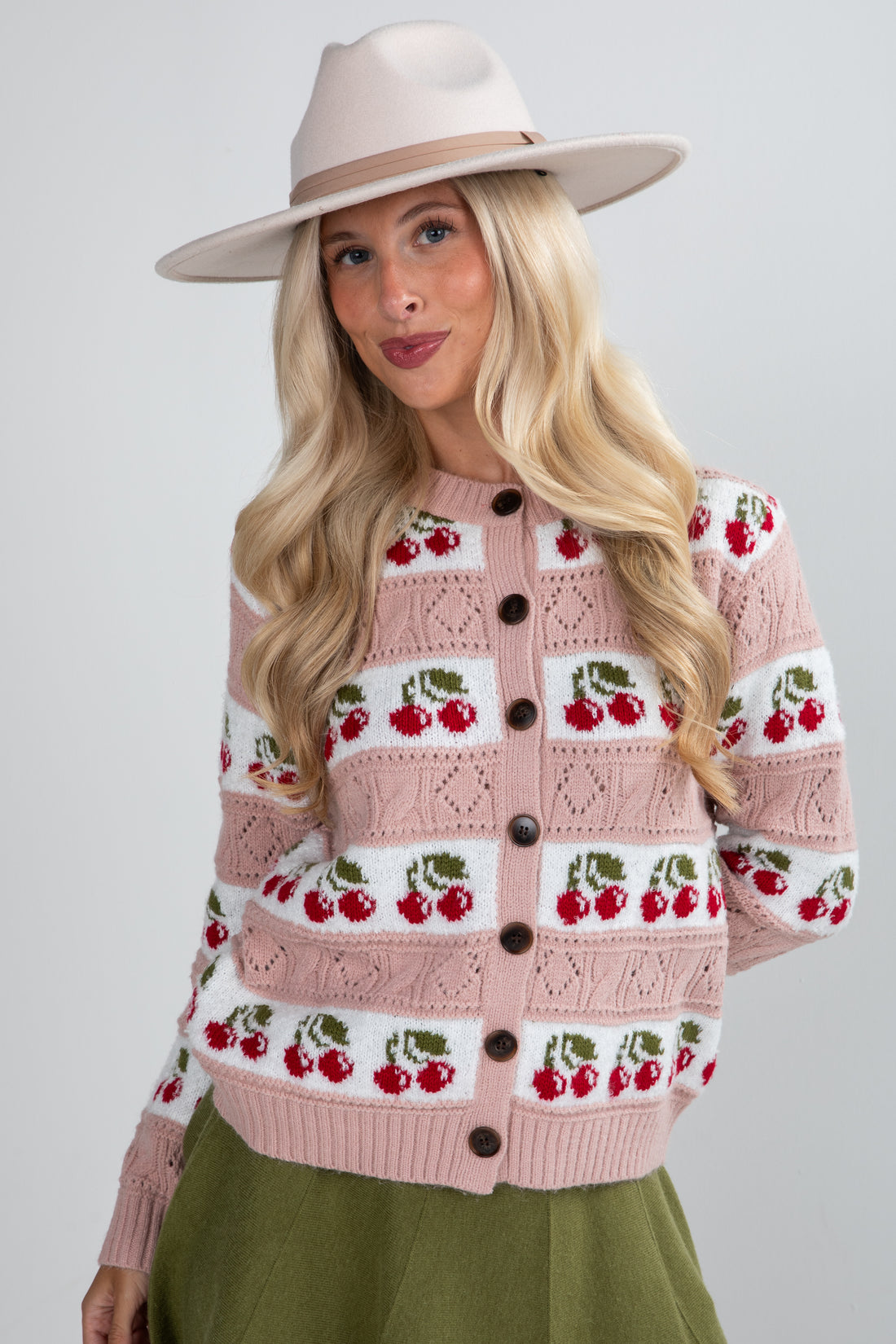 Woman wearing a pink cardigan with cherry pattern and a beige hat on a light gray background